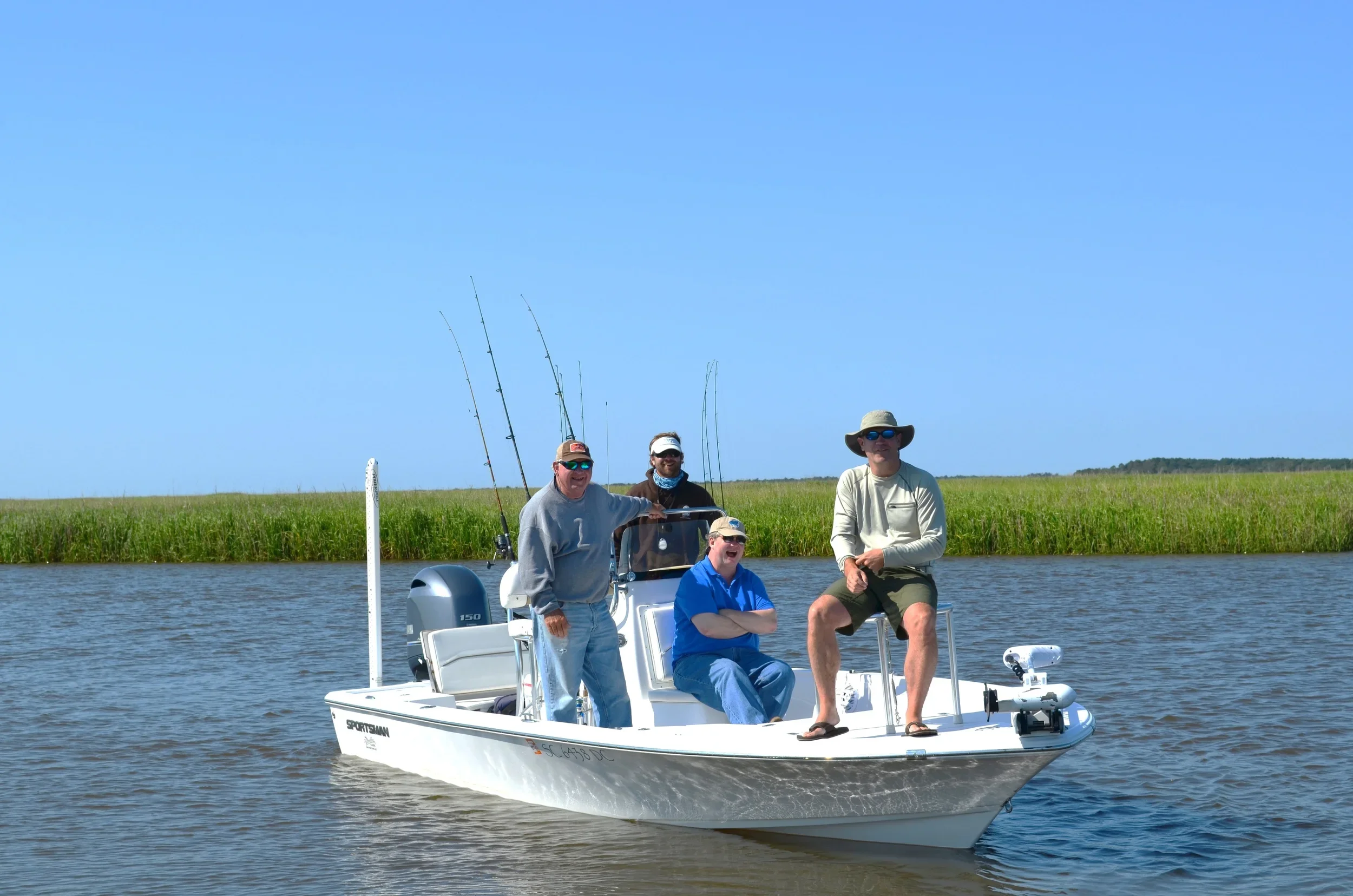 Four men on a small fishing boat on a lake, with a grassy shoreline and blue sky in the background. The men are dressed casually and are smiling or looking at the camera, with fishing rods attached to the boat.