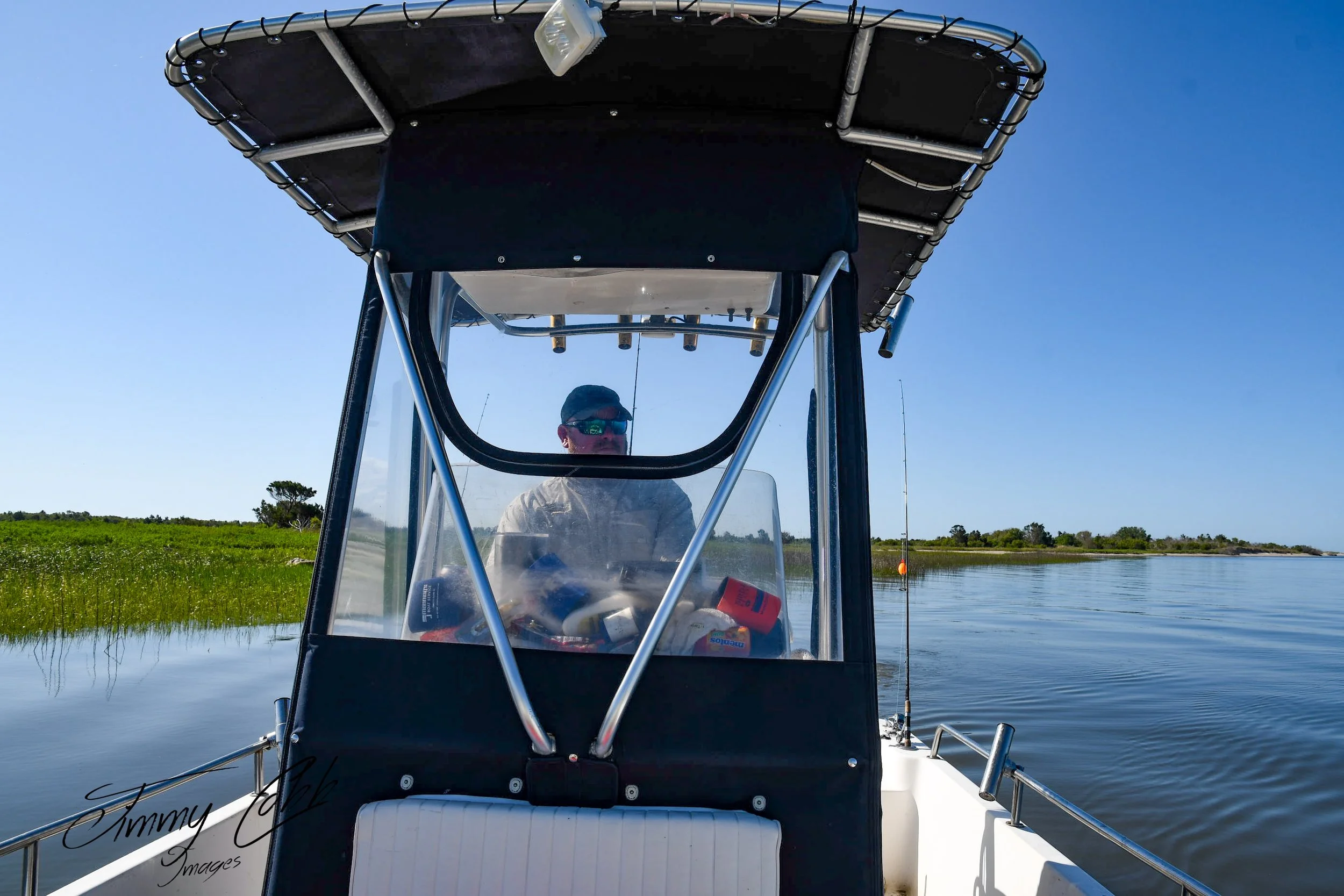 A person wearing sunglasses and a cap, sitting inside a boat with a canopy, on a calm waterway with grassy banks and trees in the background, under a clear blue sky.