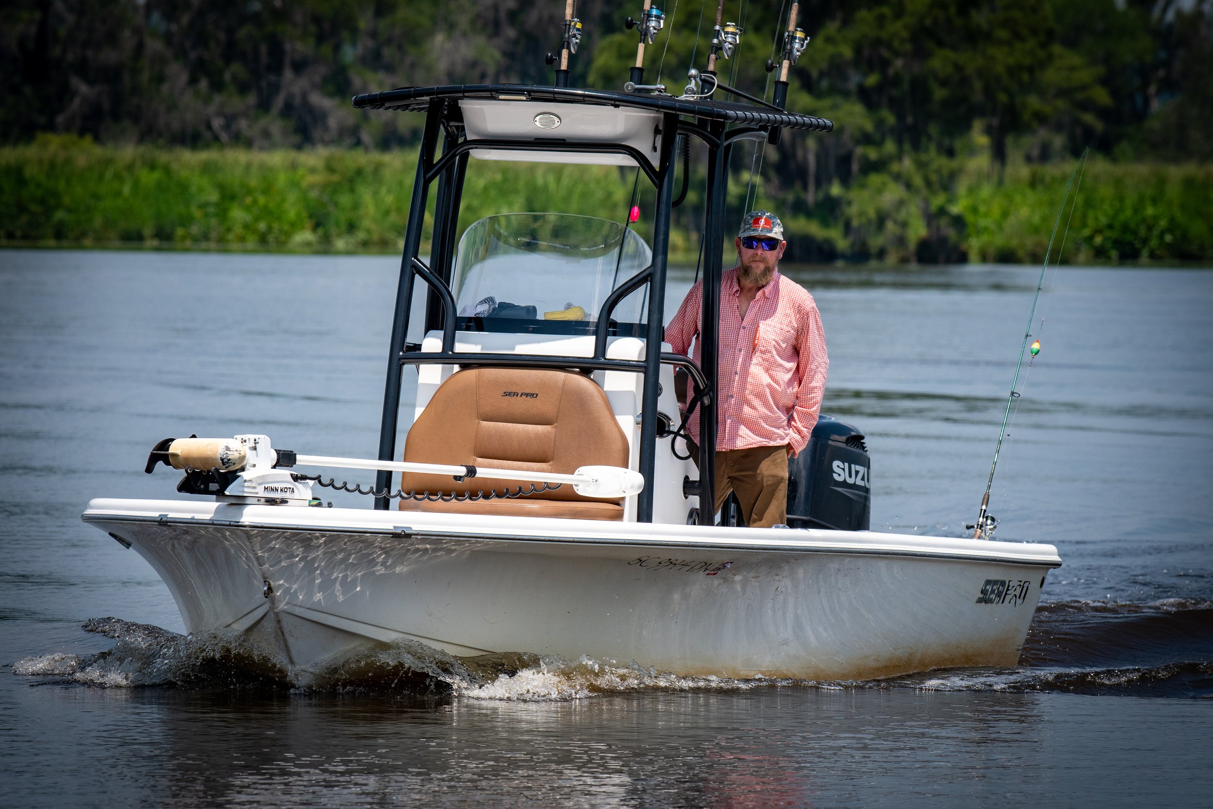 A man with a beard and sunglasses is on a small white motorboat on a body of water, with a fishing rod in hand, surrounded by green trees.
