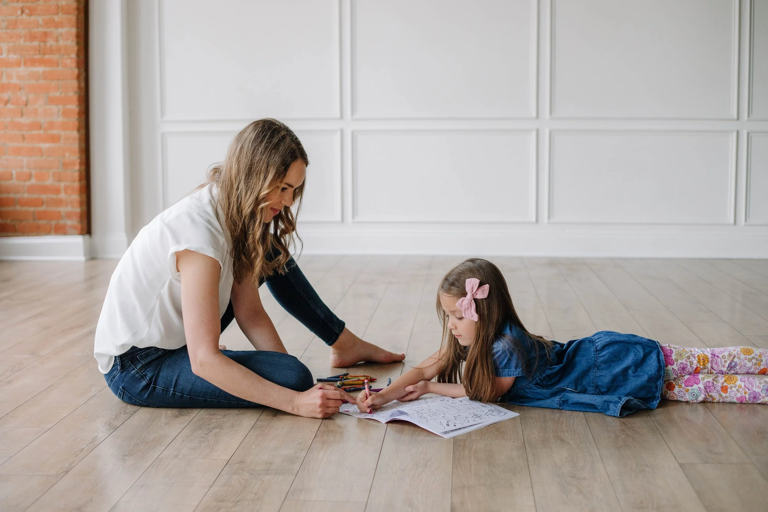 A woman and a young girl are sitting on a wooden floor indoors. The woman is helping the girl with drawing or coloring in a sketchbook. The girl is lying on her stomach with a pink bow in her hair, and there are colored pencils scattered nearby. The room has white walls with decorative panels and a brick wall on the left.