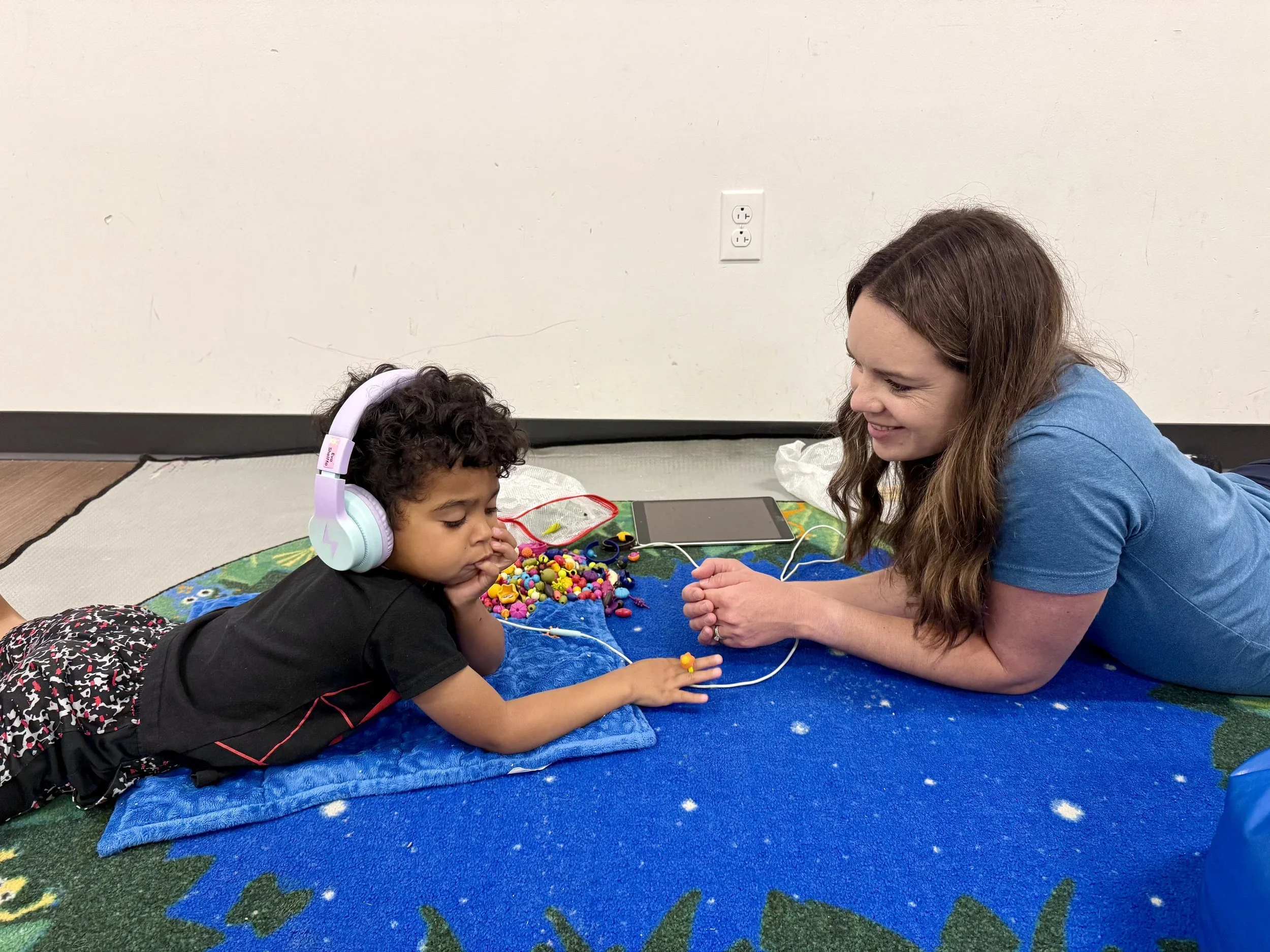 An occupational therapist playing ont he floor with a young child wearing head phones, working on sensory processing and emotional regulation during occupational therapy for kids in Dallas, TX