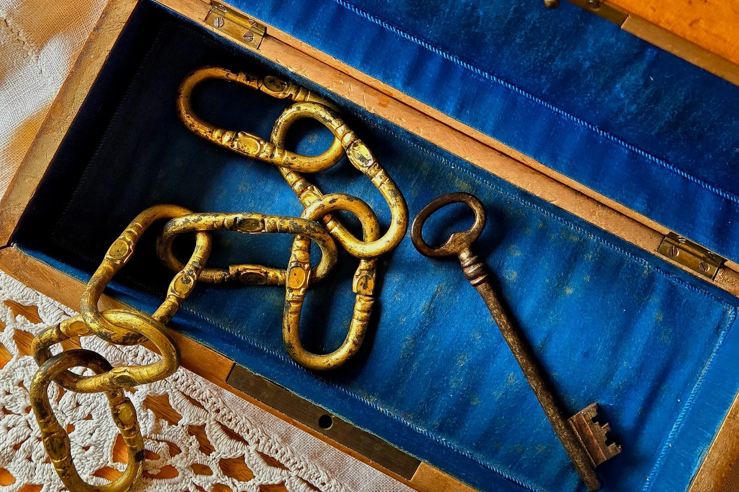 An open vintage wooden box lined with blue satin, containing an antique gilded chain and skeleton key, placed on a white lace tablecloth.