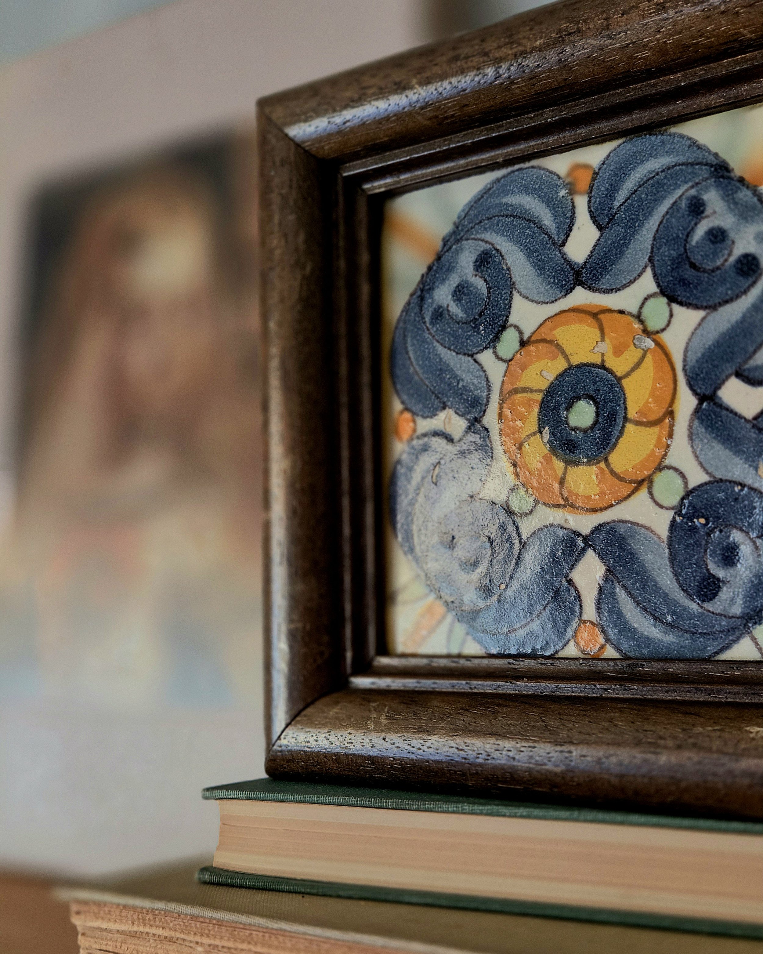 A framed ceramic tile with colorful abstract floral design, partially visible, on a stack of books with a green hardcover at the bottom.