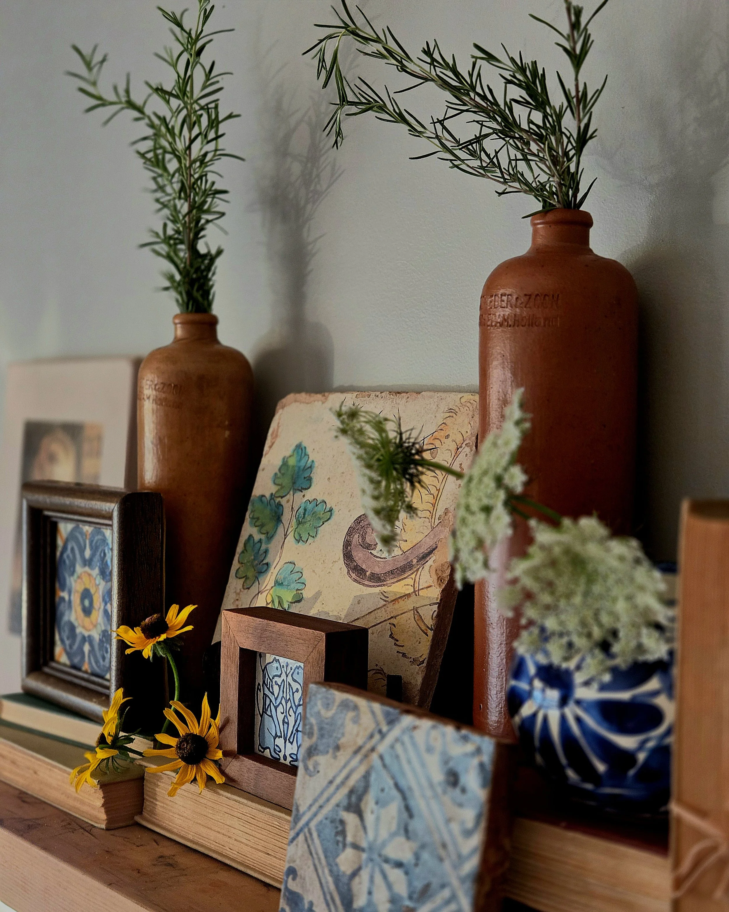 Close-up of decorative ceramic bottles with green sprigs, framed tiles, and yellow flowers on a wooden surface. Background shows a blurred wall and part of a painting or photograph.