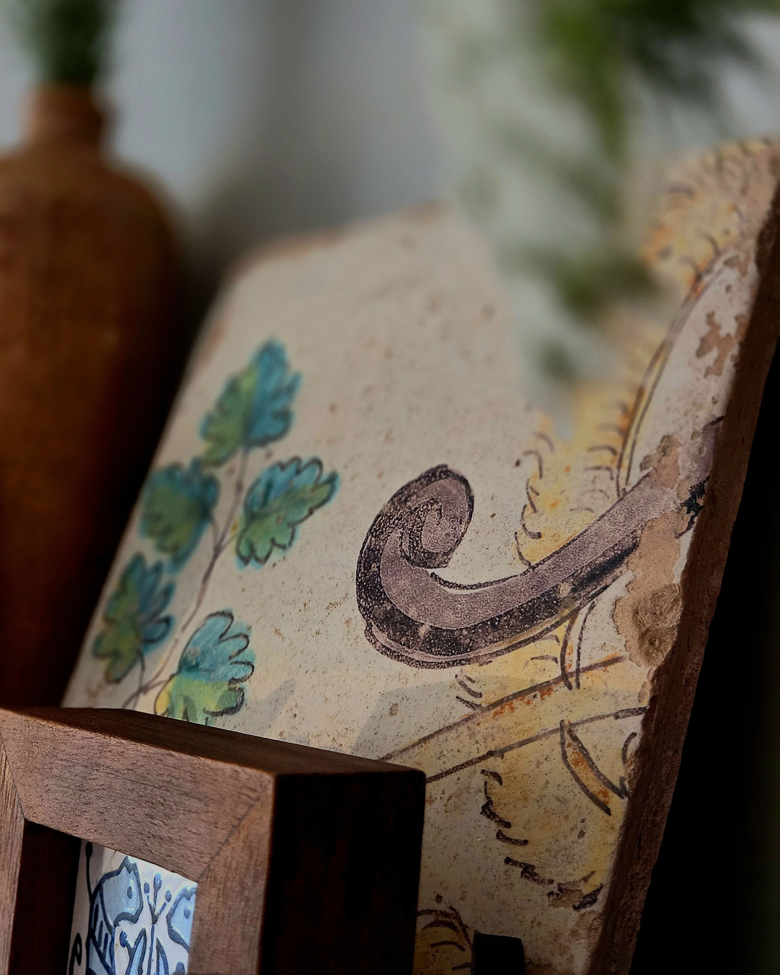 Close-up of a colorful antique tile with a curved horn shape, oak leaves, and acorns, with a blurred piece of rosemary in the foreground.