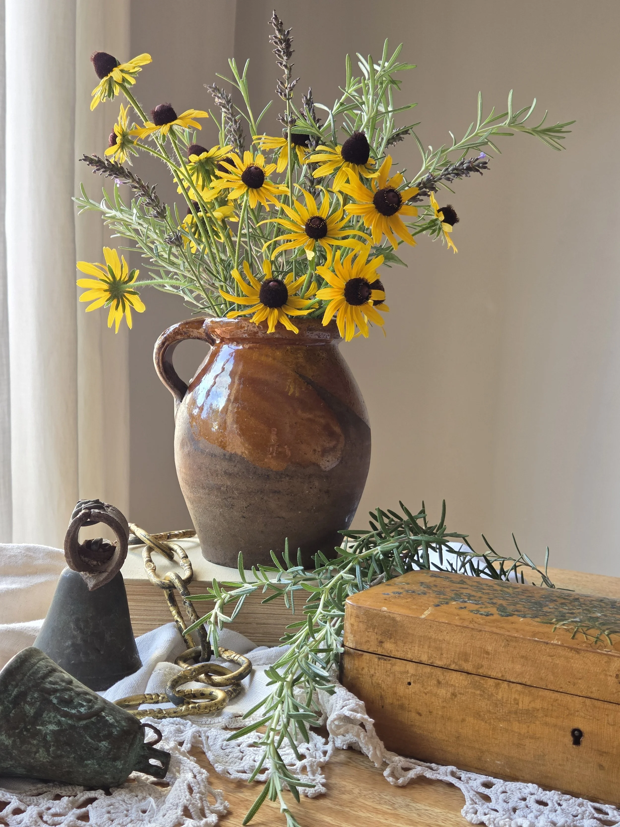 A rustic ceramic pitcher with yellow black-eyed Susan flowers, purple lavendar, and green foliage, displayed on a wooden surface with antique items including a brass chain, a verdigris green bell, and a wooden box, set against a plain wall.