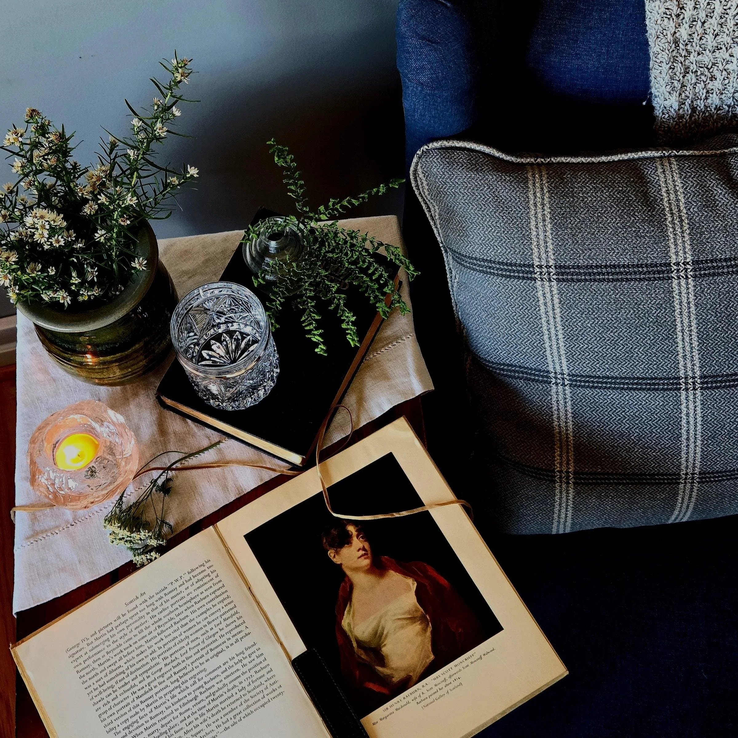 Open book featuring a portrait of a woman and text, with decorative candles and green plants in vases on a table.