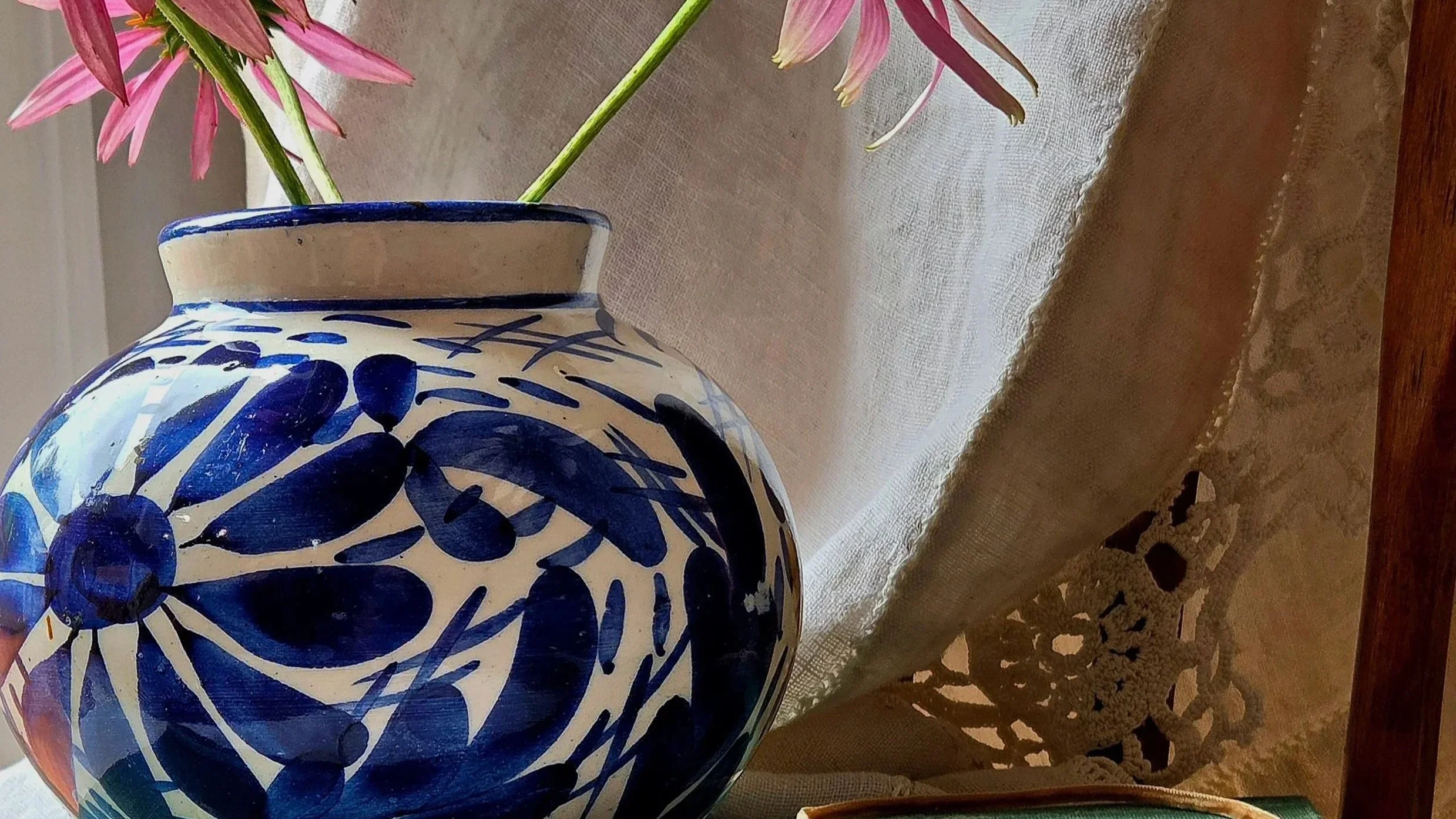 A ceramic vase with blue floral patterns holding pink flowers on a wooden surface, with a lace curtain and textured beige wall in the background.