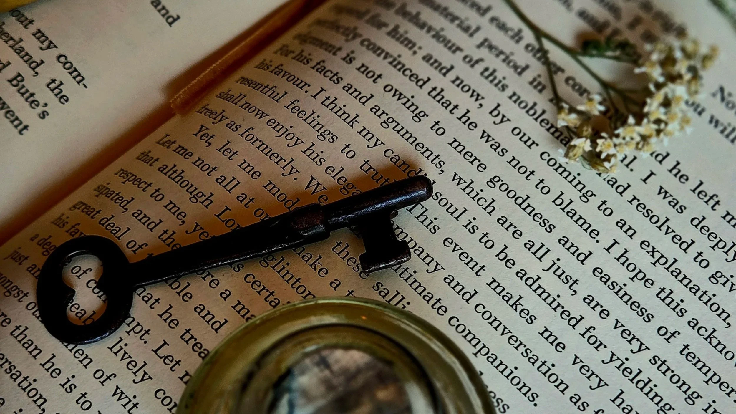 Close-up of an open book with text, a small bunch of tiny white flowers, a black metal key, and a small glass jar container on top.