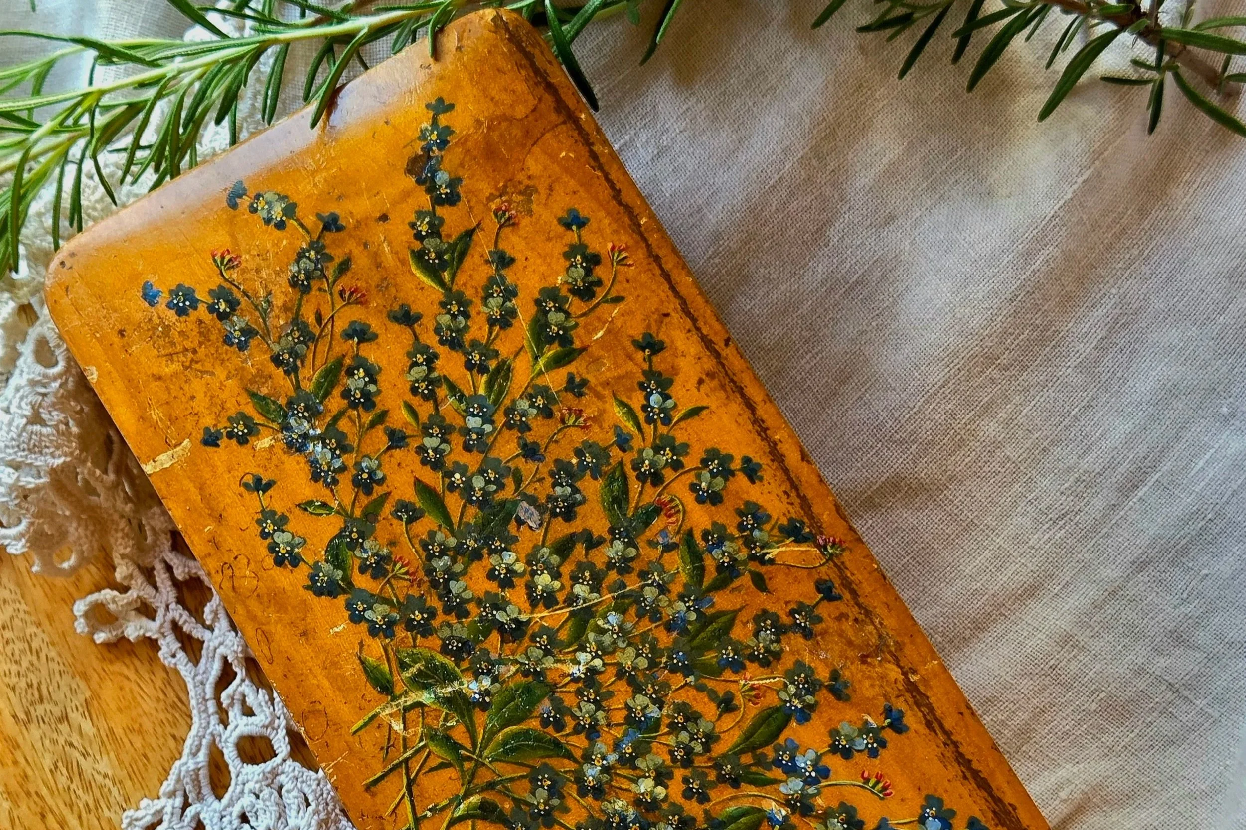 A vintage wooden box with a painted floral design, surrounded by green leaves and white lace on a wooden surface.