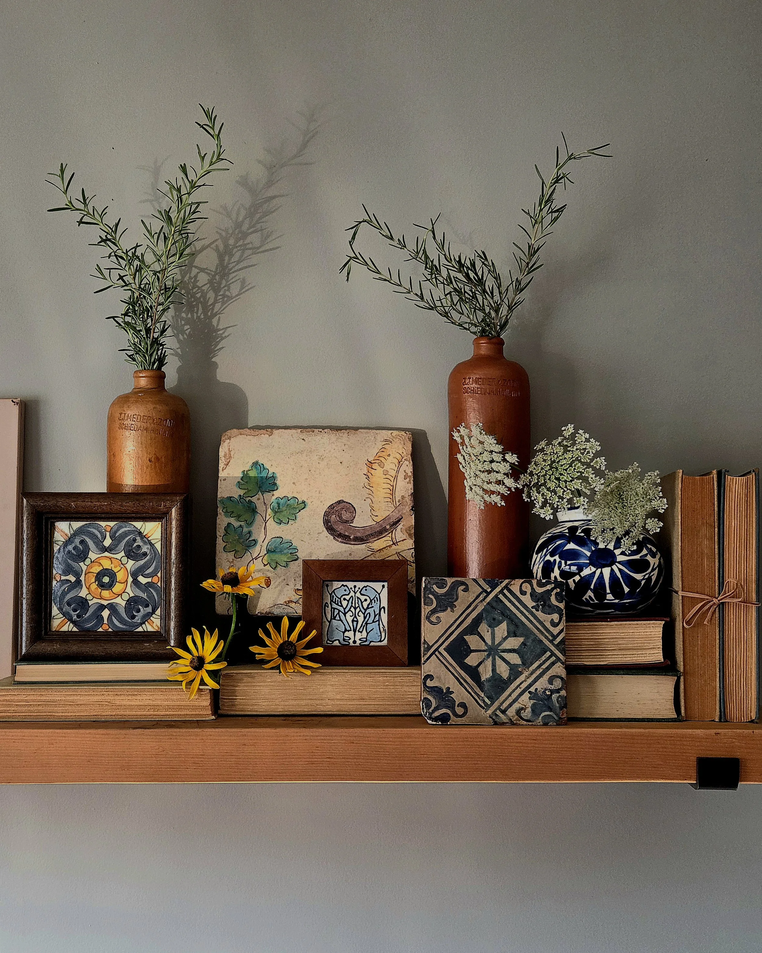 Arrangement of decorative items on a wooden shelf, including two tall ceramic bottles holding green foliage, small framed tile artwork, ceramic vase with flowers, and patterned tiles.