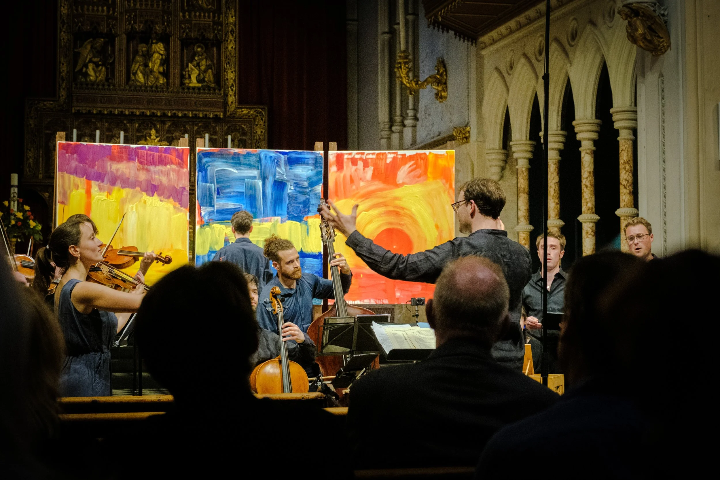 Chamber orchestra performing in an ornate church with colorful abstract paintings behind the musicians