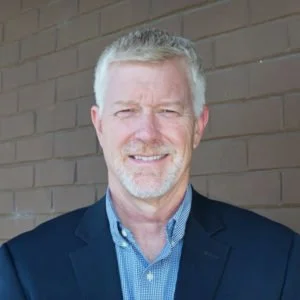 A smiling middle-aged man with gray hair and beard, wearing a navy blazer over a blue collared shirt, standing in front of a brick wall.