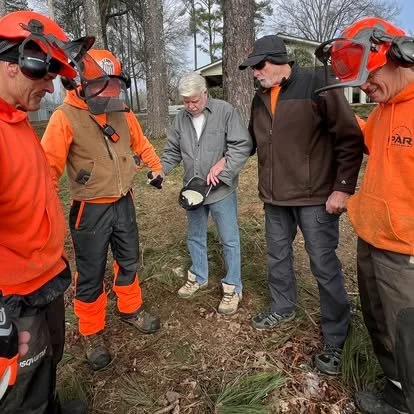 Group of five people outdoors, engaged in a discussion. Three are wearing orange and black protective gear with helmets and goggles, while one person is in a gray jacket and another in a black jacket with a cap. They are standing on a wooded area with trees and a building in the background.