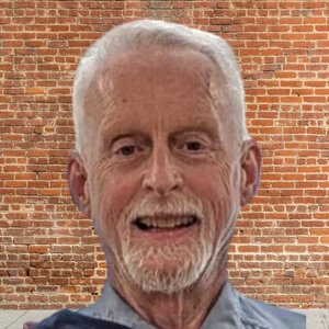 A smiling elderly man with white hair and a beard, standing in front of a brick wall.