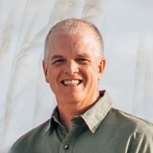 Smiling man with short gray hair wearing a light green collared shirt, outdoors with a cloudy sky background