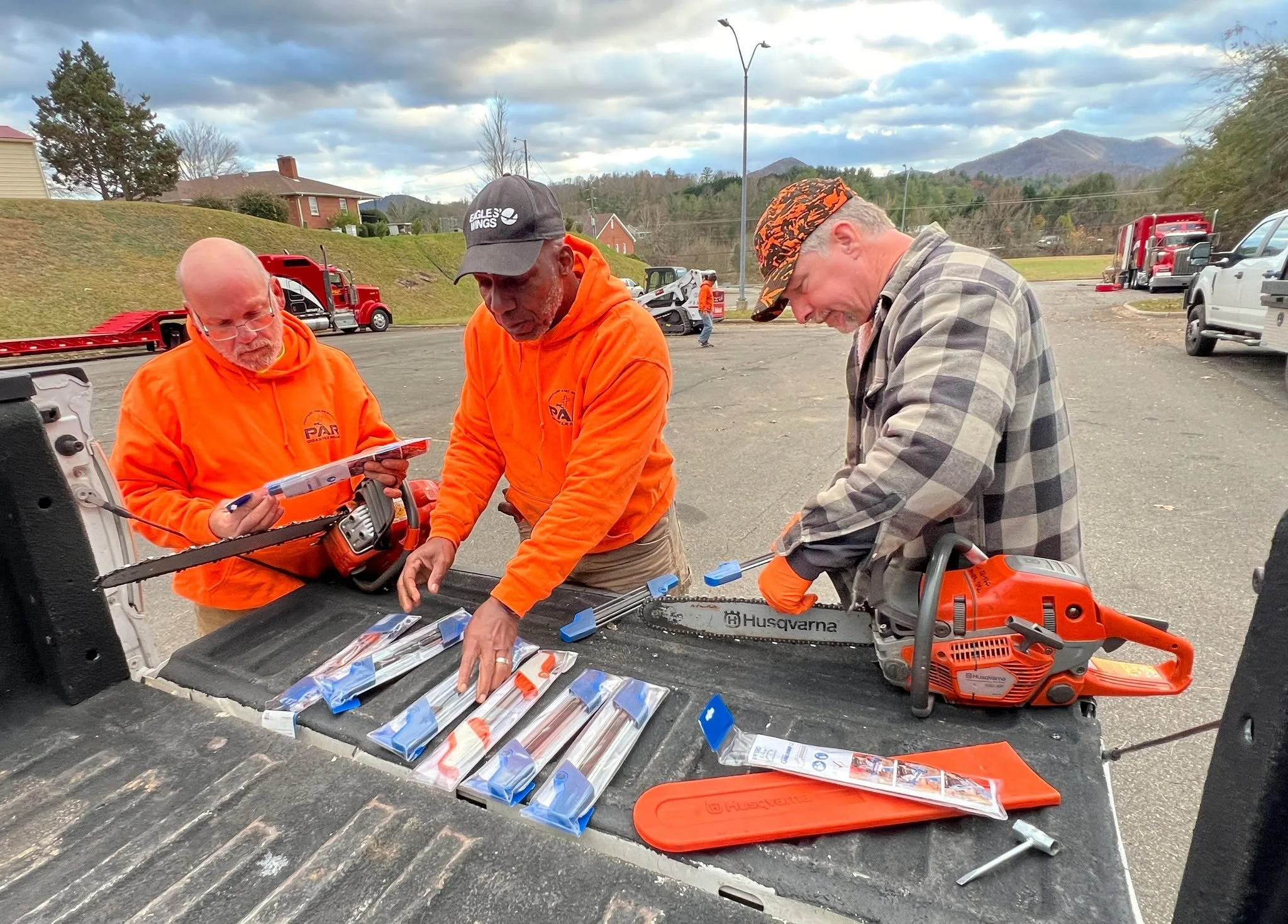 Three men gather around a work table with chainsaw parts in an outdoor parking lot with trucks and mountains in the background. One man wears an orange hoodie, another a baseball cap, and the third a plaid shirt and a patterned cap.