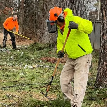 Two people clearing debris in a wooded area, one wearing a bright yellow jacket, orange helmet, and face shield using a tool, and the other wearing an orange jacket working with a rake.