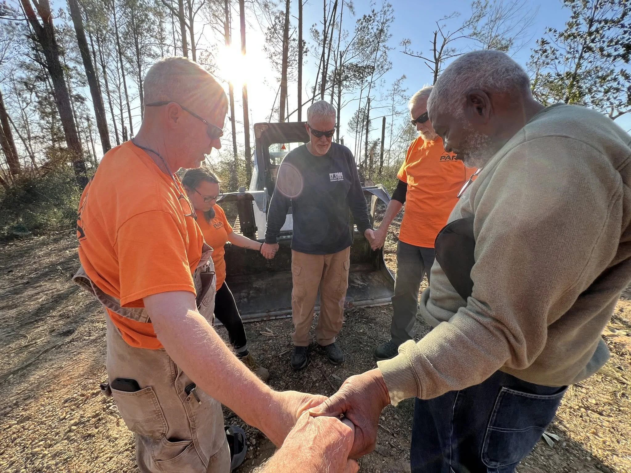 A group of people standing outdoors in a circle, holding hands, with a wooded area and the sun shining in the background.