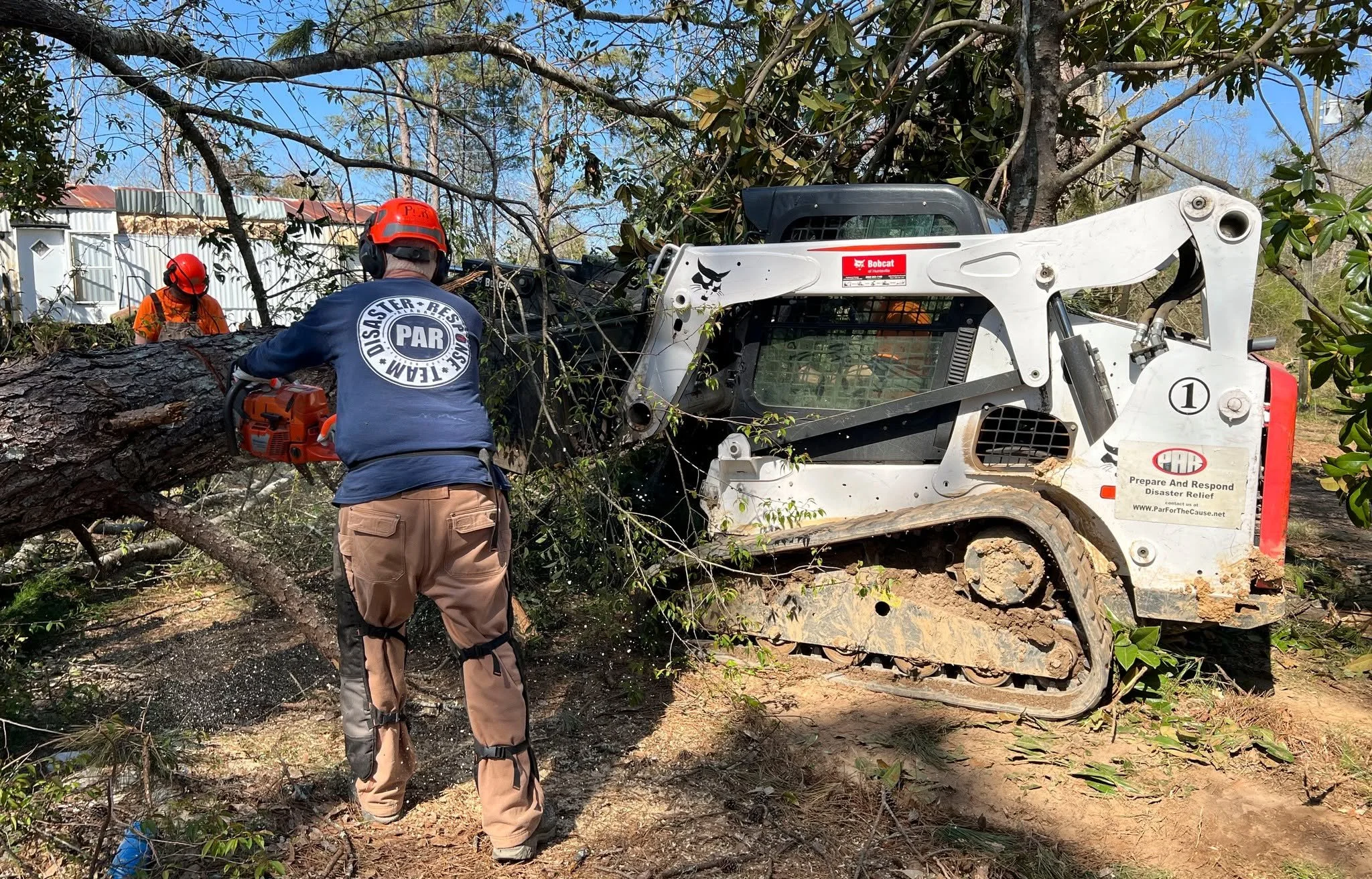 Workers utilizing a Bobcat compact track loader to remove a fallen tree in a backyard, surrounded by trees and a fence, on a clear day.