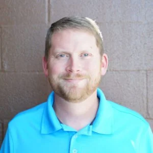 A man with short blond hair, a beard, and light skin wearing a bright blue polo shirt, standing against a tan brick wall, smiling.