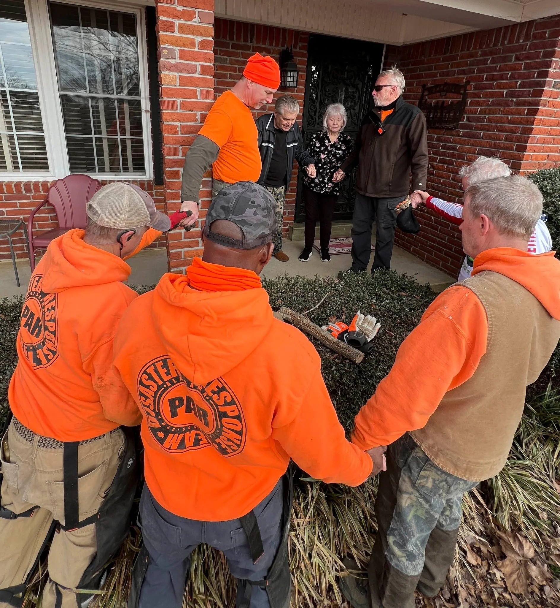 A group of people standing closely together outside a brick house, with some wearing bright orange hoodies, one with a matching orange hat, and others dressed in dark or camo clothing, possibly engaged in a shared activity or prayer.