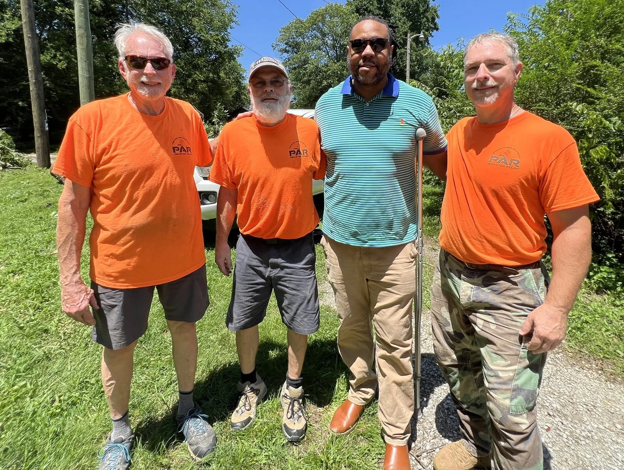 Four men standing outdoors on a sunny day, two wearing orange shirts with a logo, one in a striped polo shirt with sunglasses, and one in camouflage shorts with a crutch nearby, surrounded by green trees and grass.