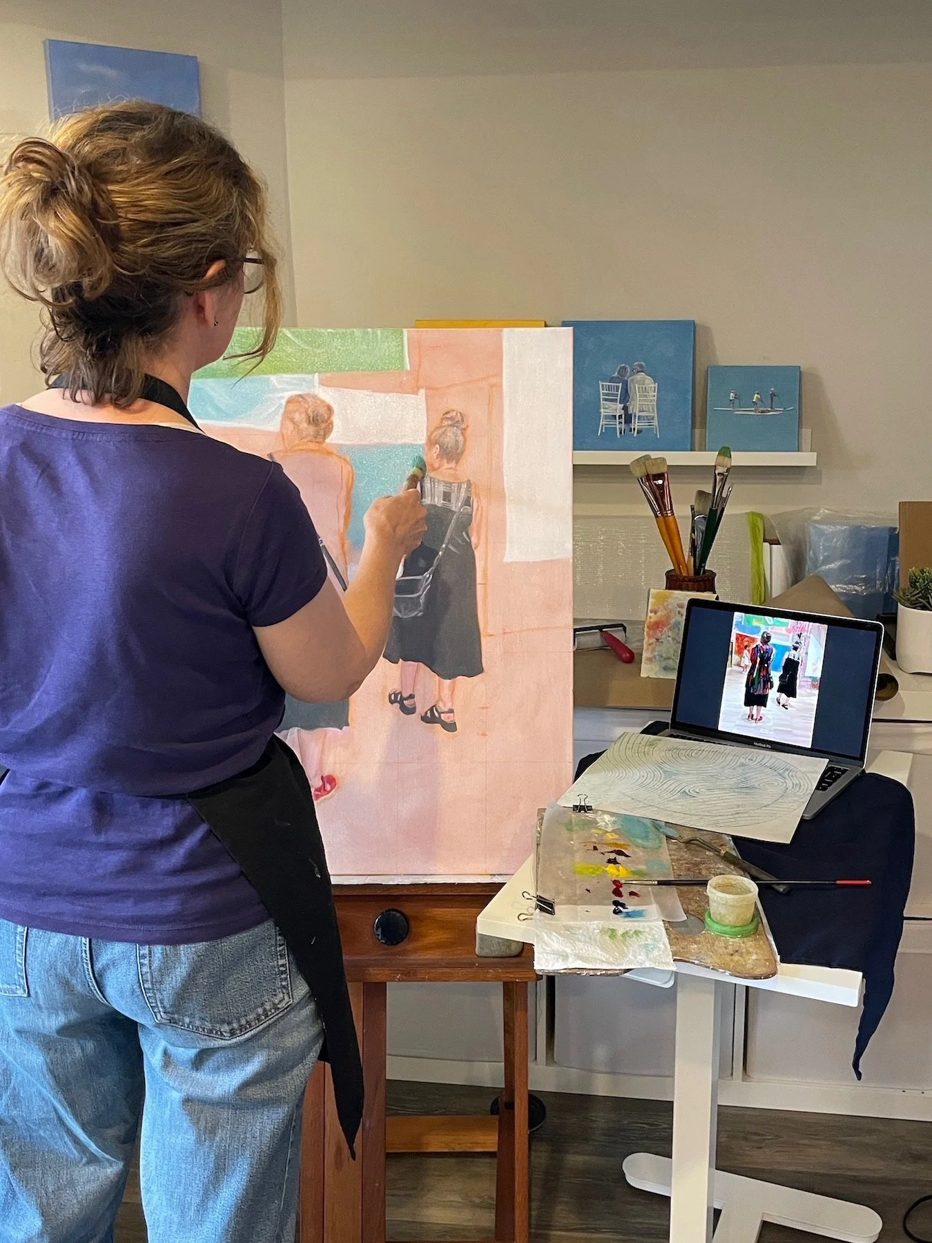 An artist painting a portrait of two women on a canvas. The artist is using brushes and the workspace includes art supplies, a laptop, and small blue paintings on the wall.