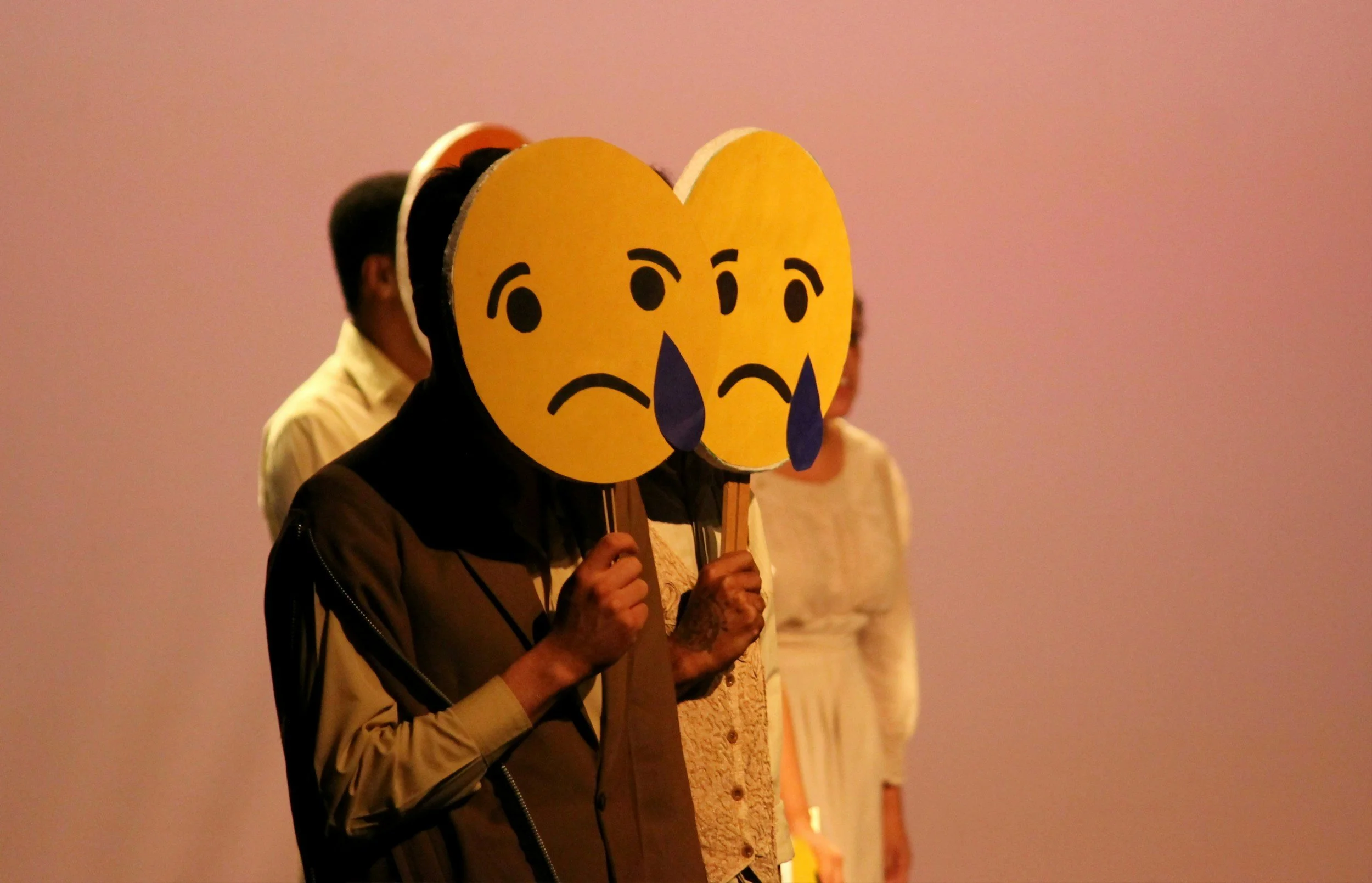People standing on a stage holding large emoji-style signs with sad, crying faces.