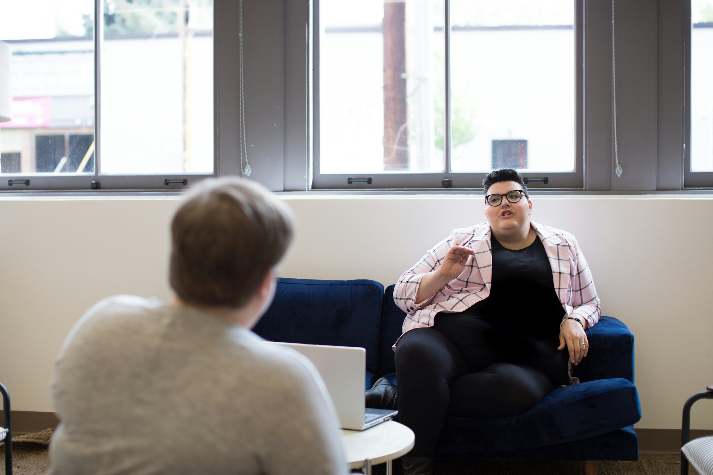 Two people are having a conversation in a room with large windows. One person is sitting on a blue couch, wearing glasses and a pink checkered blazer, gesturing with their hand. The other person, with short brown hair, is sitting at a white table facing away from the camera.