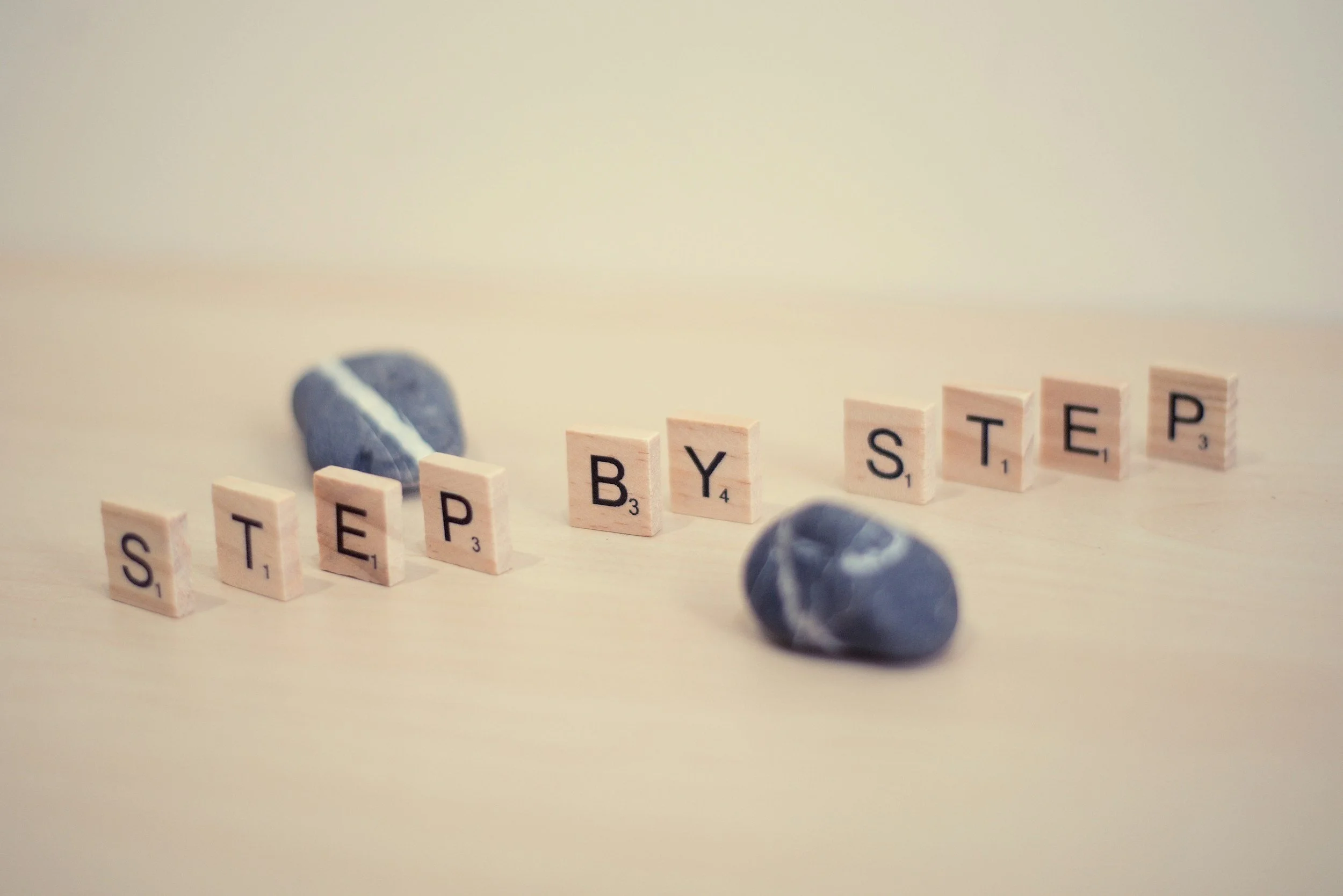 Scrabble tiles spelling out 'STEP BY STEP' on a beige surface with two blue marbles in the foreground and a third partially visible marble behind the tiles.