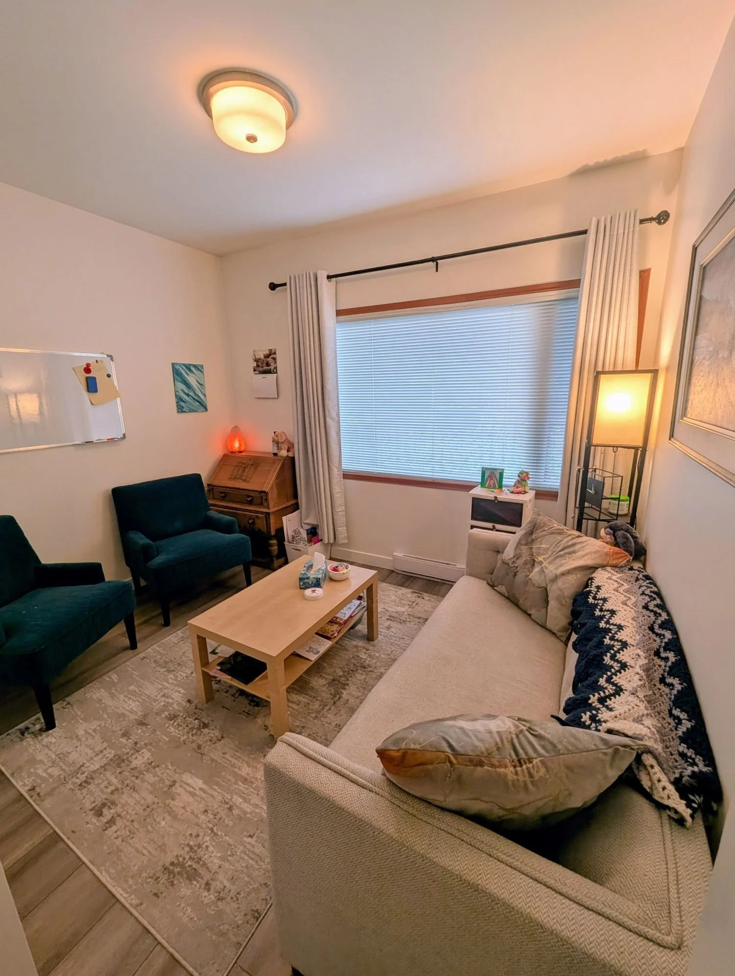 Kim Sholinder’s counselling office in Victoria, BC. Room with beige sofa, green armchairs, wooden coffee table, white rug, large window with blinds and white curtains, decorative items, and a ceiling light fixture.