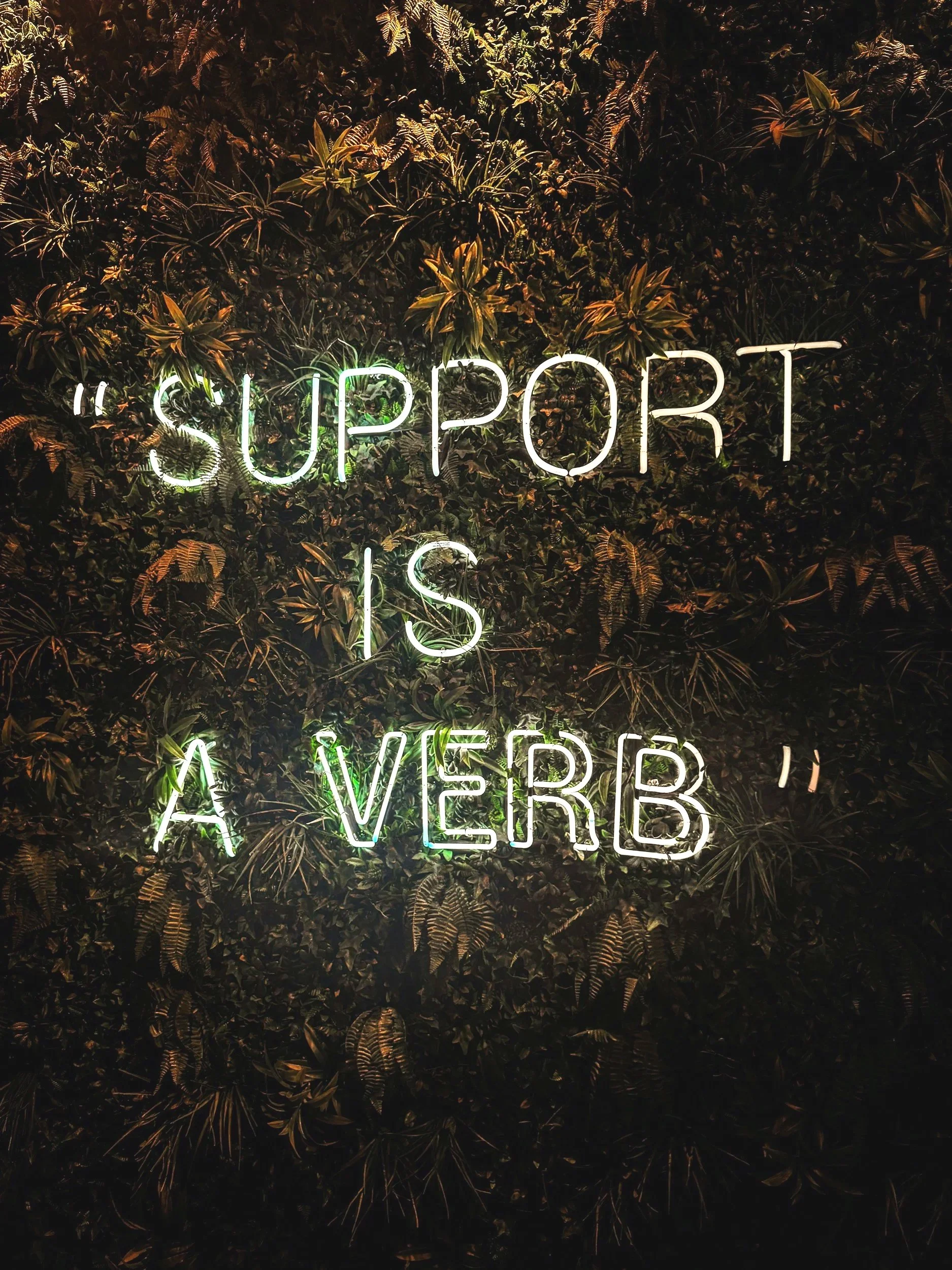 Green neon sign on a living wall that says "Support is a verb".