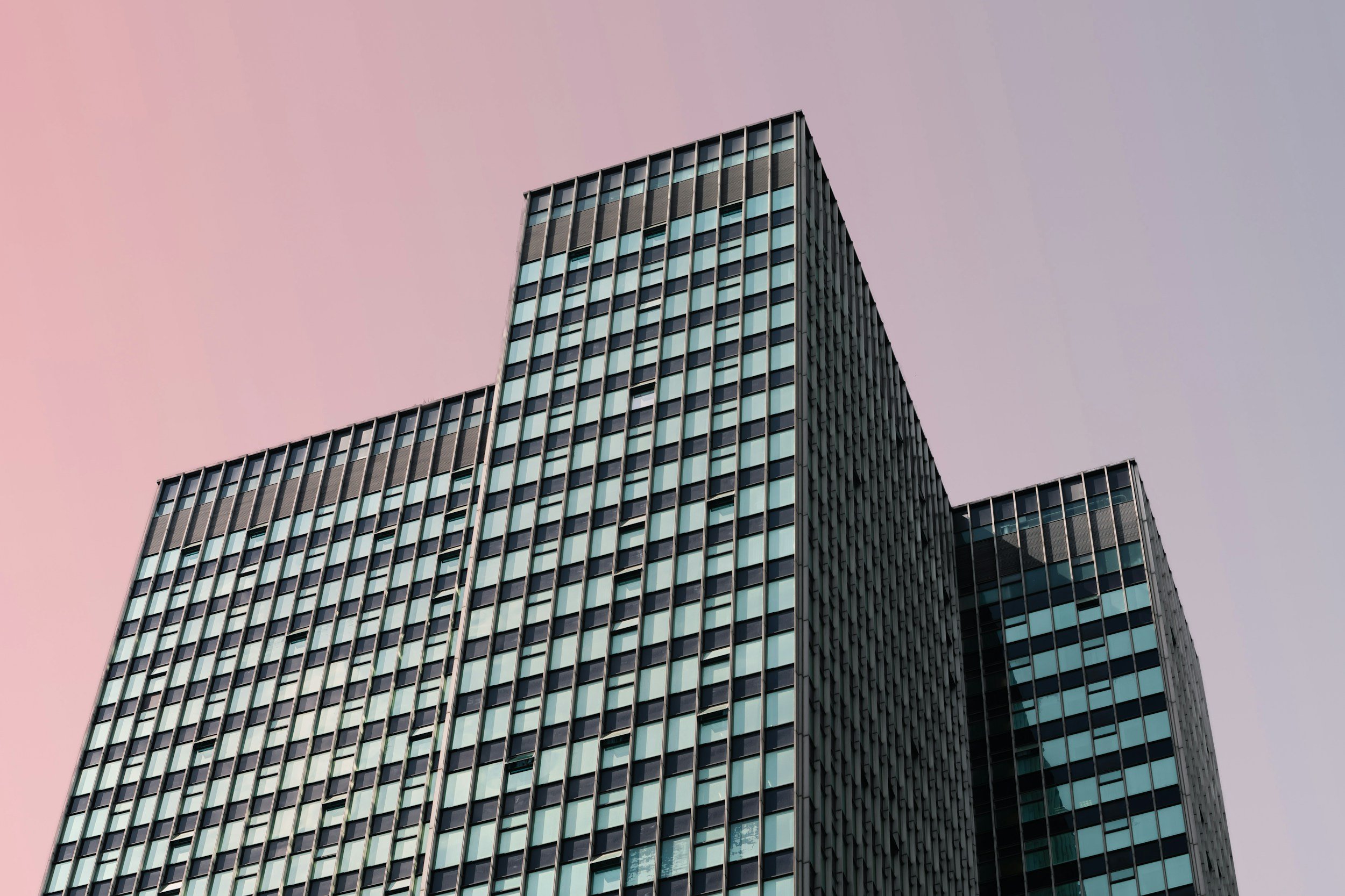 A modern glass and steel skyscraper with numerous windows against a pastel-colored sky, possibly at dawn or dusk.