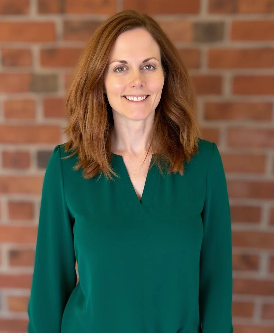 A smiling woman with shoulder-length red hair wearing a green blouse standing in front of a brick wall.