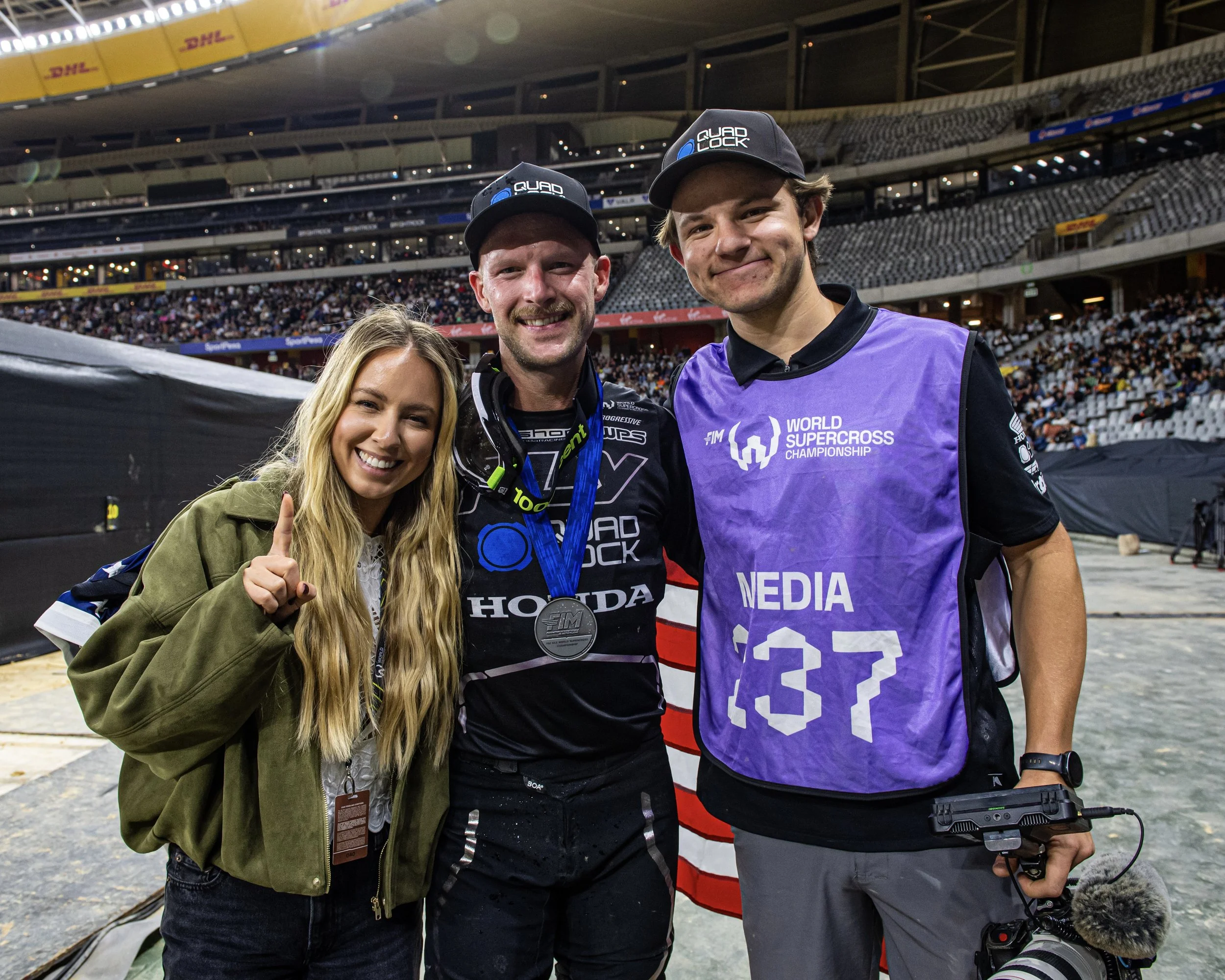 Three people smiling at a motorsports event, two men wearing black caps and racing gear, and a woman with blonde hair wearing an olive-green jacket, at a stadium with a crowd, banners, and track visible in the background.