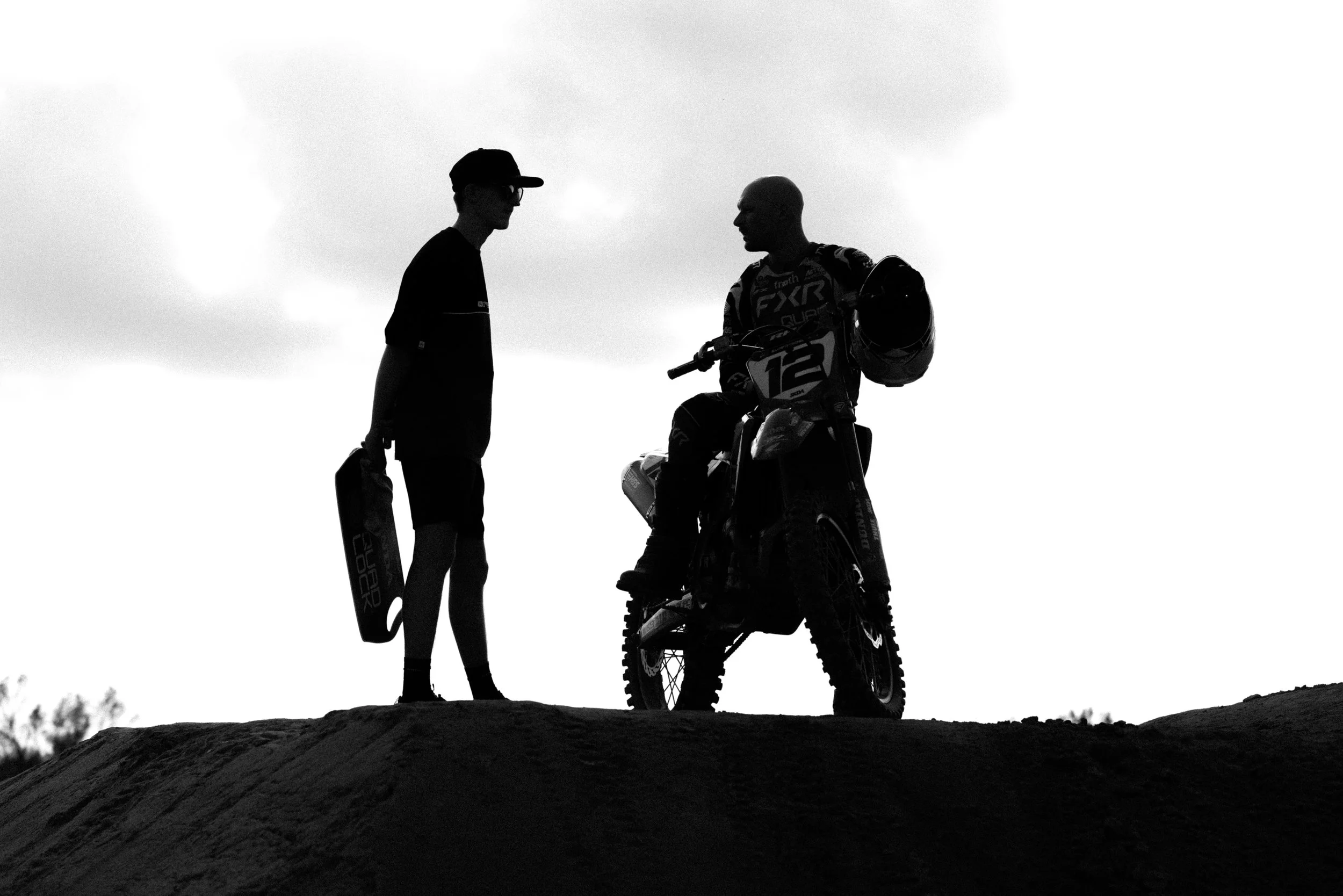 Silhouettes of two people, one on a motocross bike and the other holding a helmet, standing on a dirt hill against a cloudy sky.