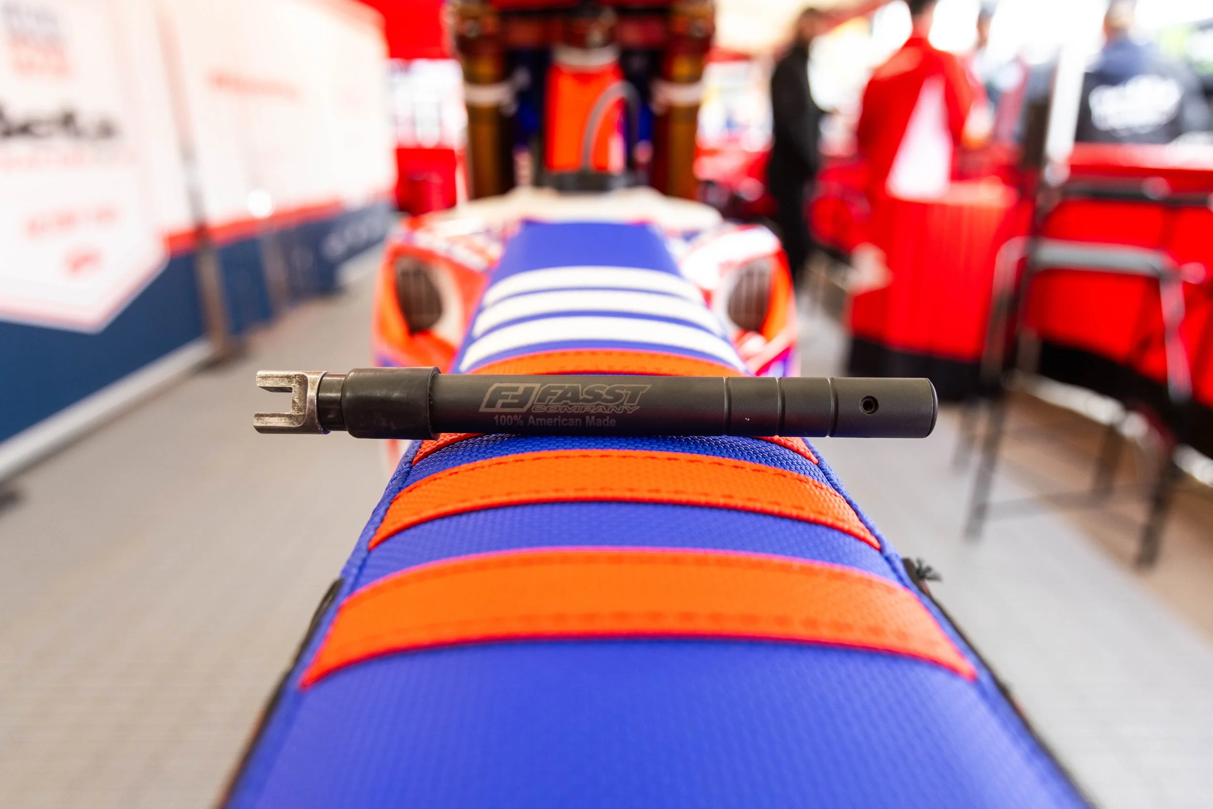 First-person view of a race car driver's foot in a racing shoe resting on the pedal, with a pen resting on the shoe, on the racing shoe. The background is blurred, showing team members and equipment in a pit area.