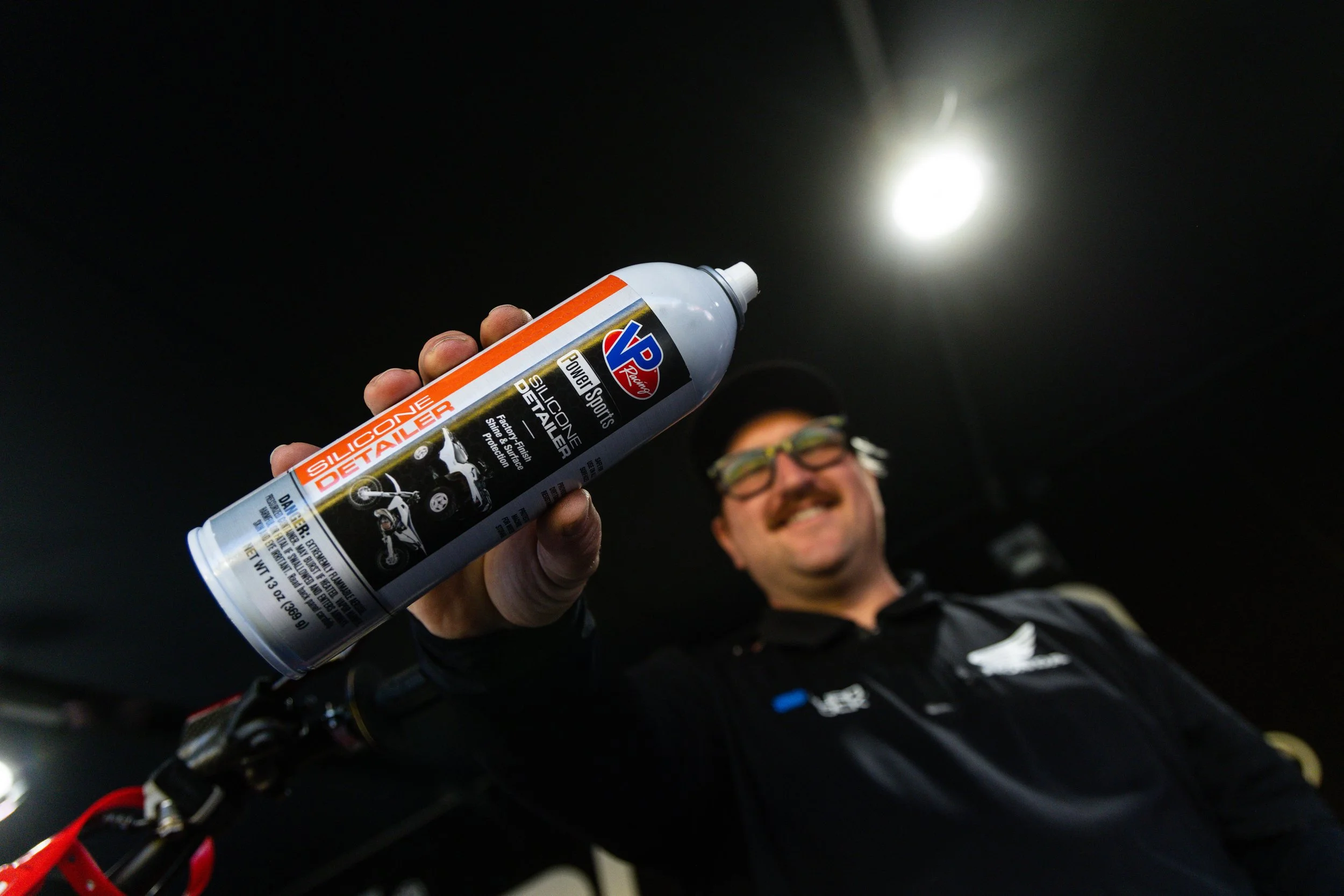 A person holding a can of VP Racing Hill Cone Silicone Detangler in front of the camera, with a bright fluorescent light overhead and a blurred person with glasses smiling in the background.