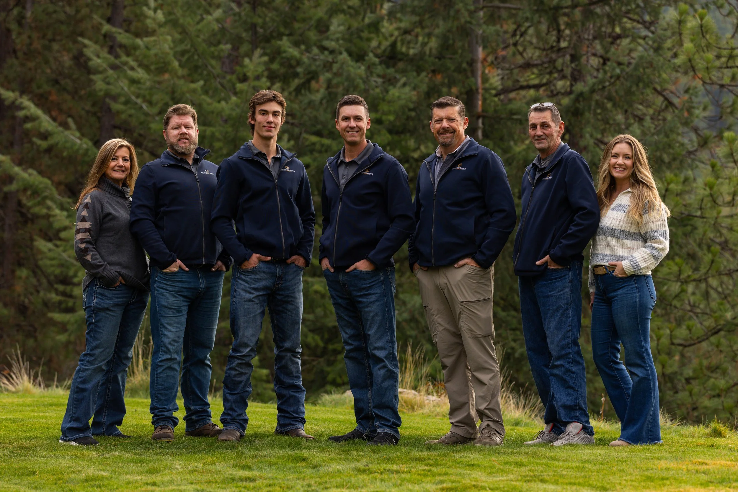 Group of seven people standing outdoors on grass in front of trees, smiling at the camera.