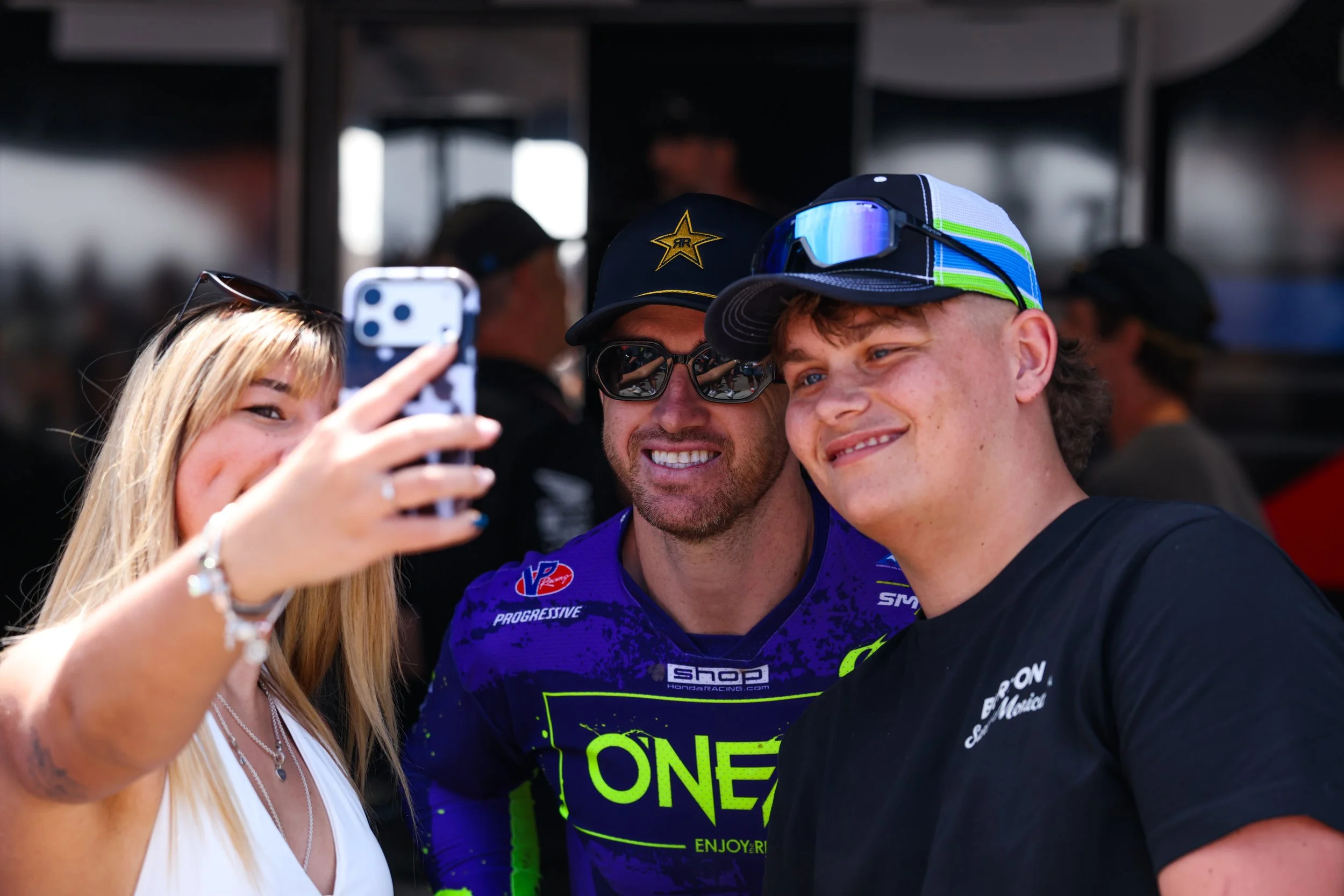 A woman with long blonde hair taking a selfie with two men, one wearing sunglasses and a hat with a star logo, and another wearing a racing cap and reflective sunglasses. The woman is smiling and holding a smartphone, while the men are smiling for the photo.