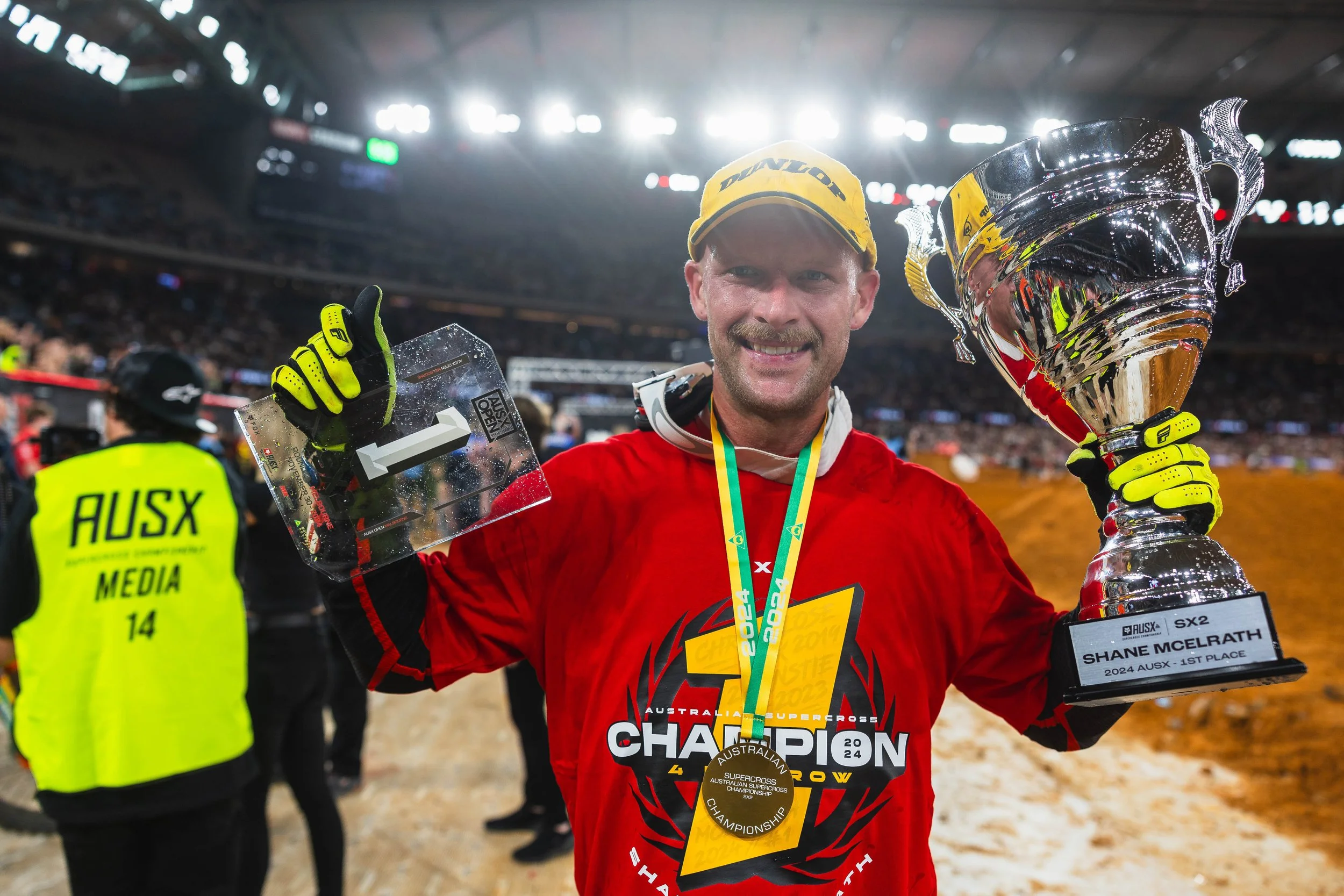 A smiling man in a red sports jersey and yellow cap holding a trophy and a plaque, celebrating winning a championship at an indoor sports arena.