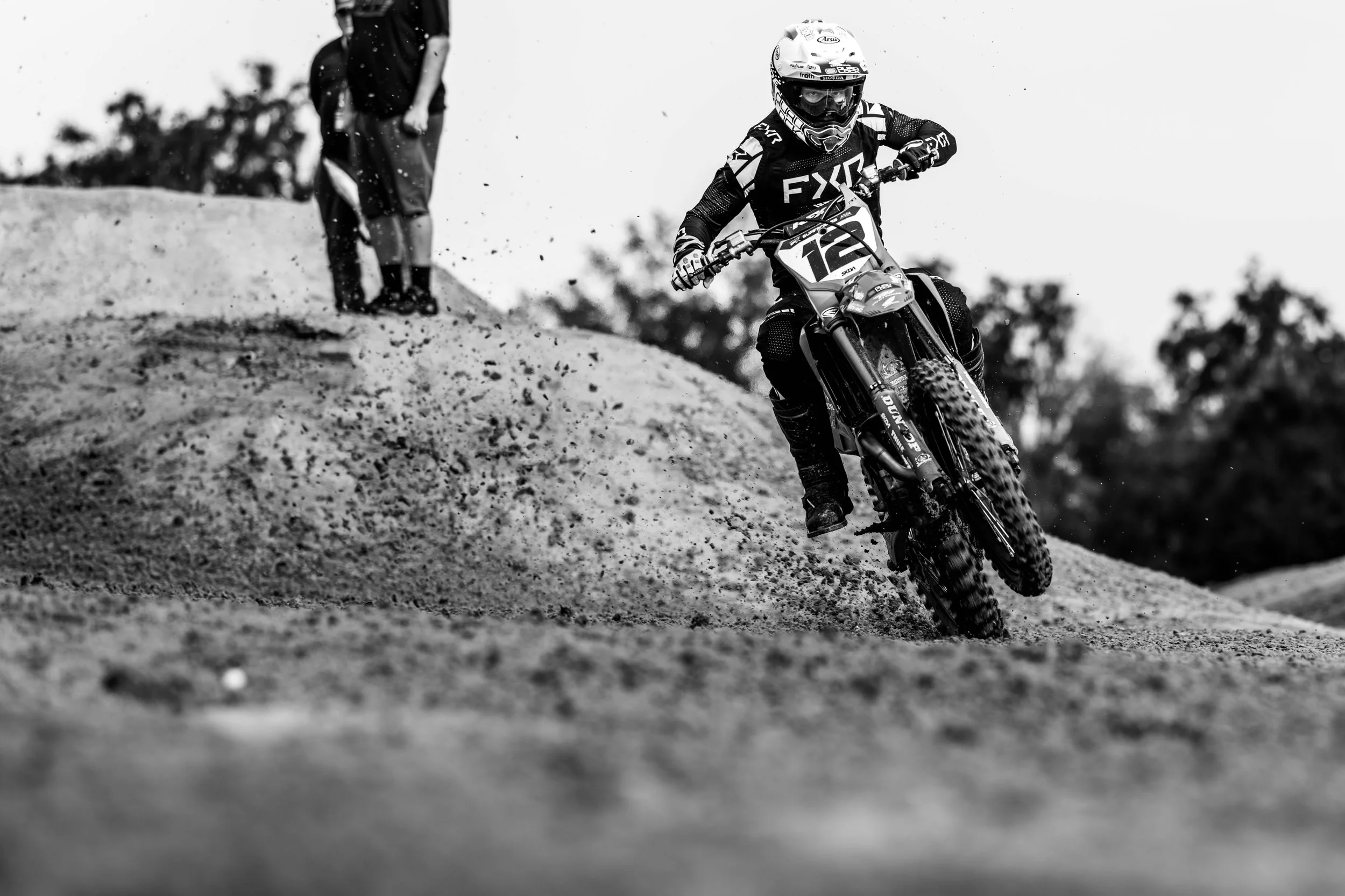 A person riding a dirt bike on a rough off-road trail, with a helmet and protective gear, kicking up dirt and gravel.