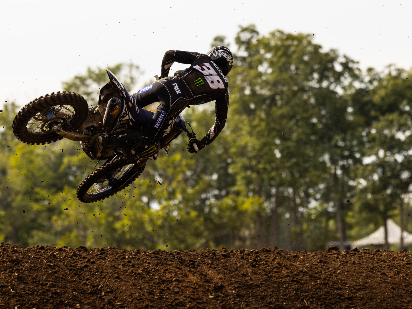 A motocross rider in black gear and helmet performing a jump on a dirt track with green trees in the background.