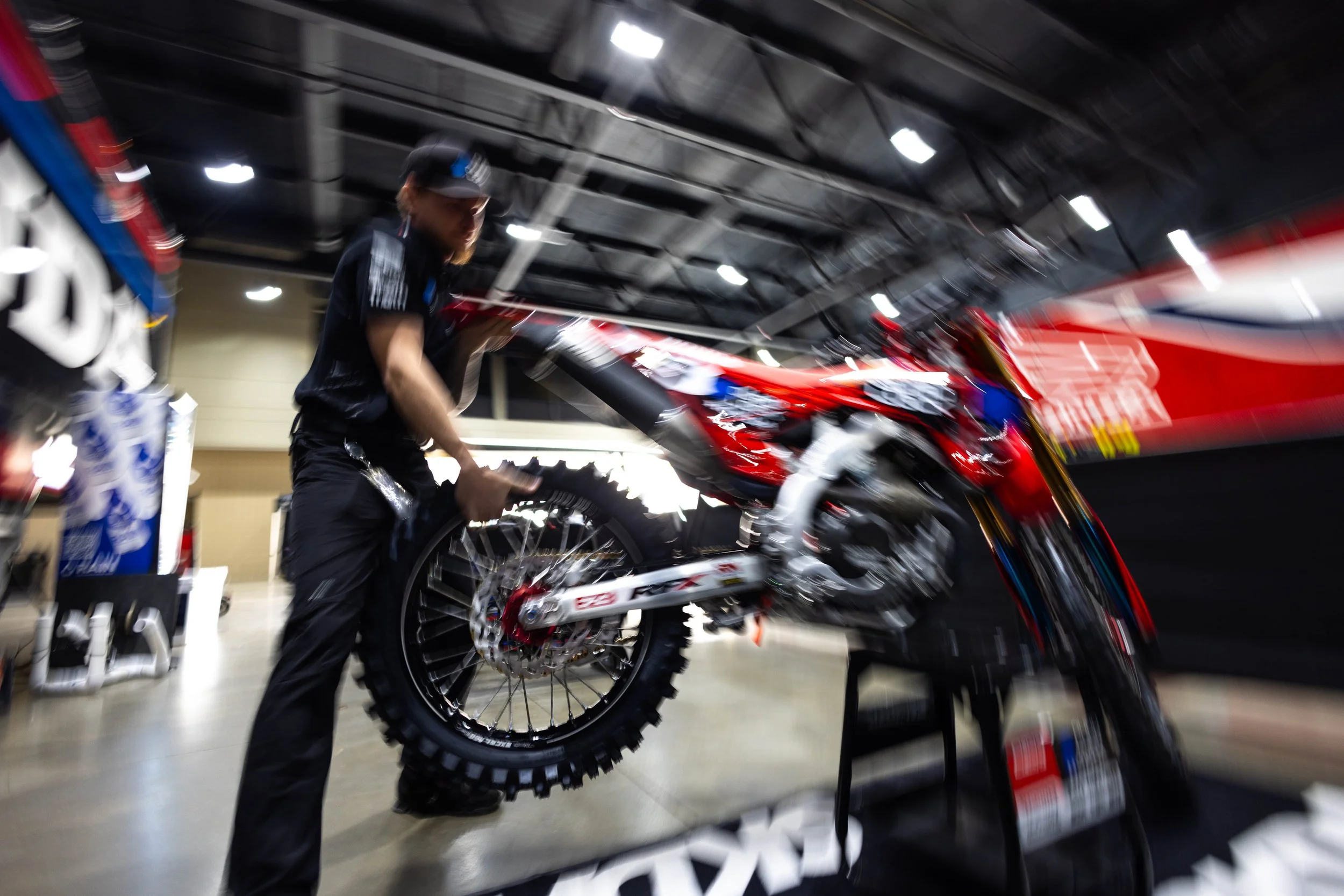 A person working on a red and black motocross bike indoors, with shelves and lighting overhead.