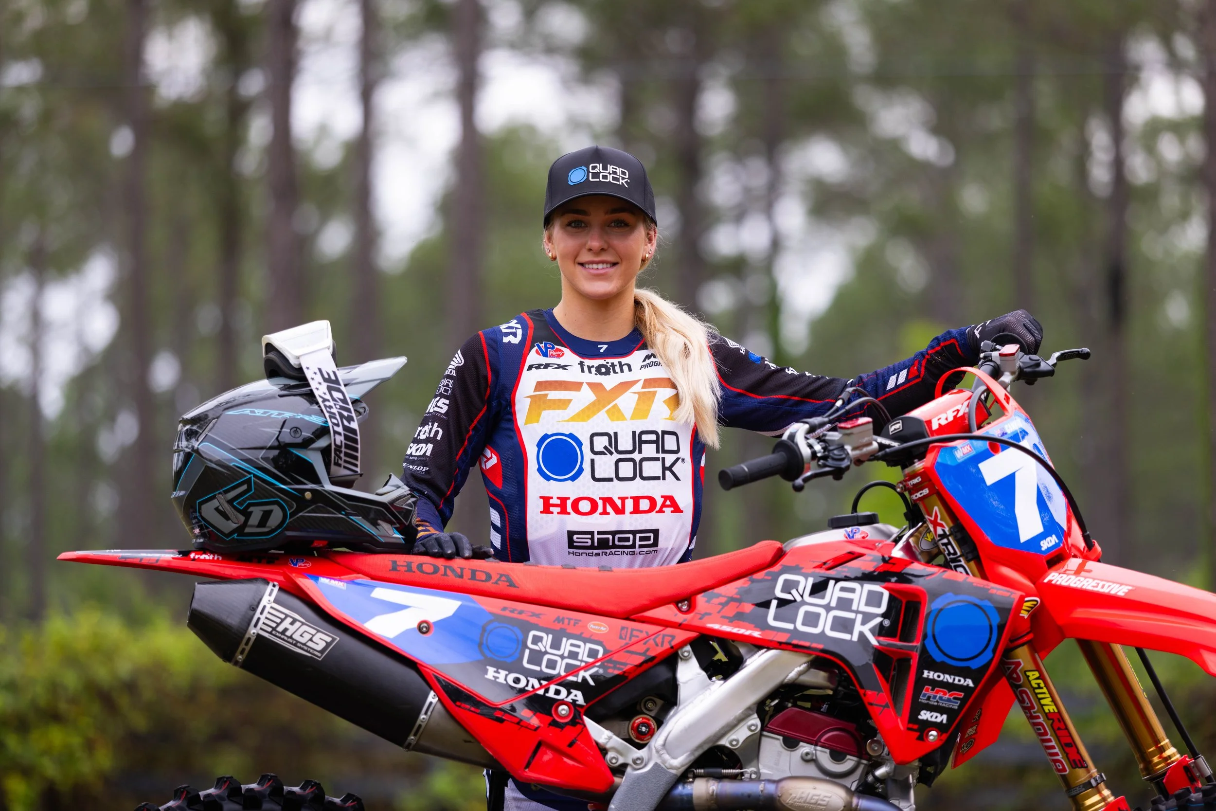 A woman in motocross gear standing beside a red Honda dirt bike, holding a helmet, with a background of trees.