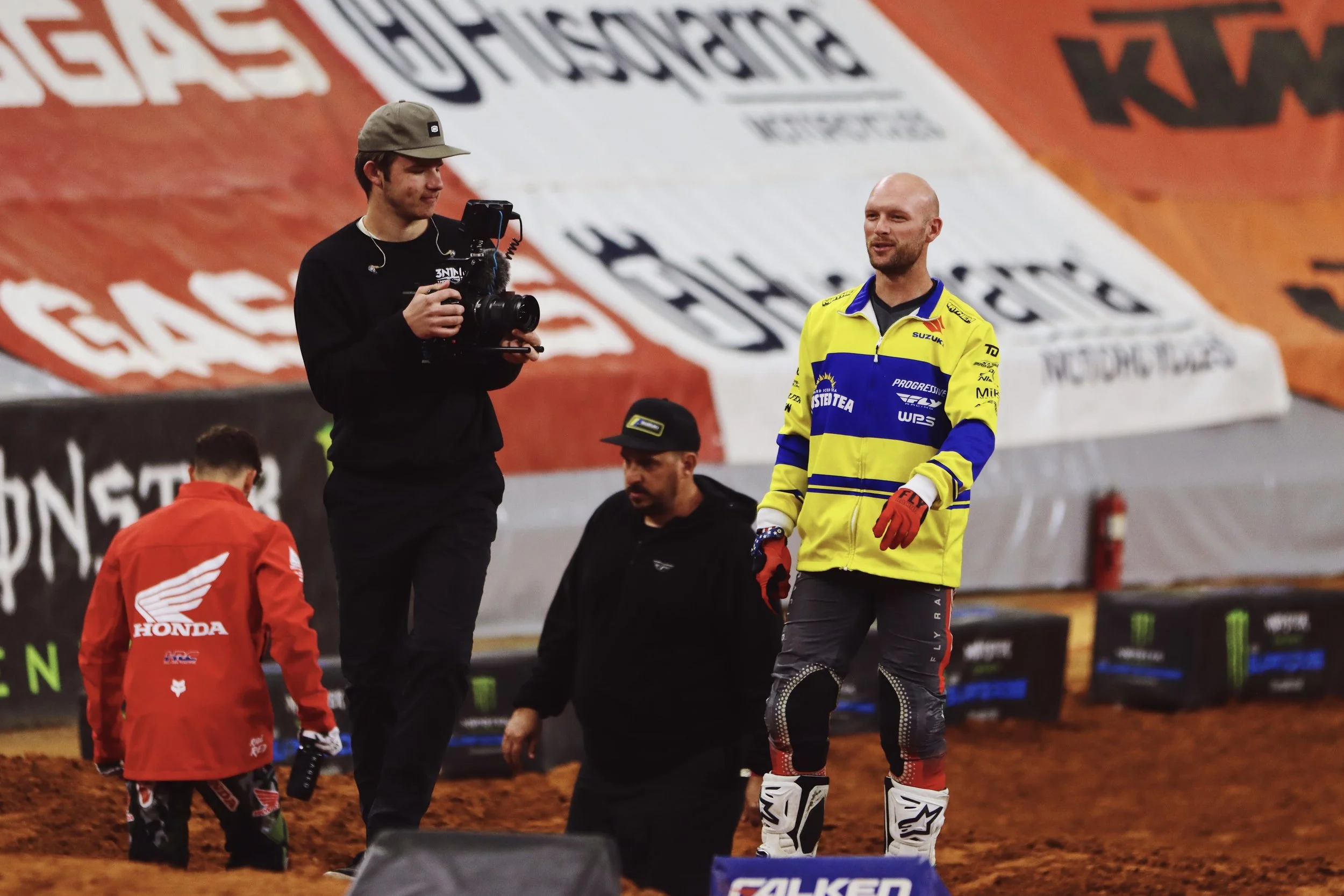 A man wearing a yellow and blue motocross jersey and red gloves stands on a dirt track, talking to a young boy in a red Honda jacket. A woman with a camera and a young man with earphones are nearby, set against a large backdrop with logos and adverti