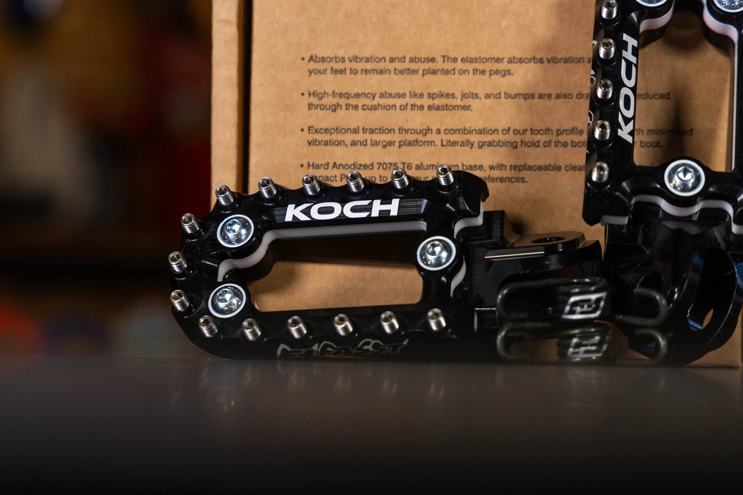Close-up of black bicycle pedals with silver screws, with a brown box in the background containing product description text.