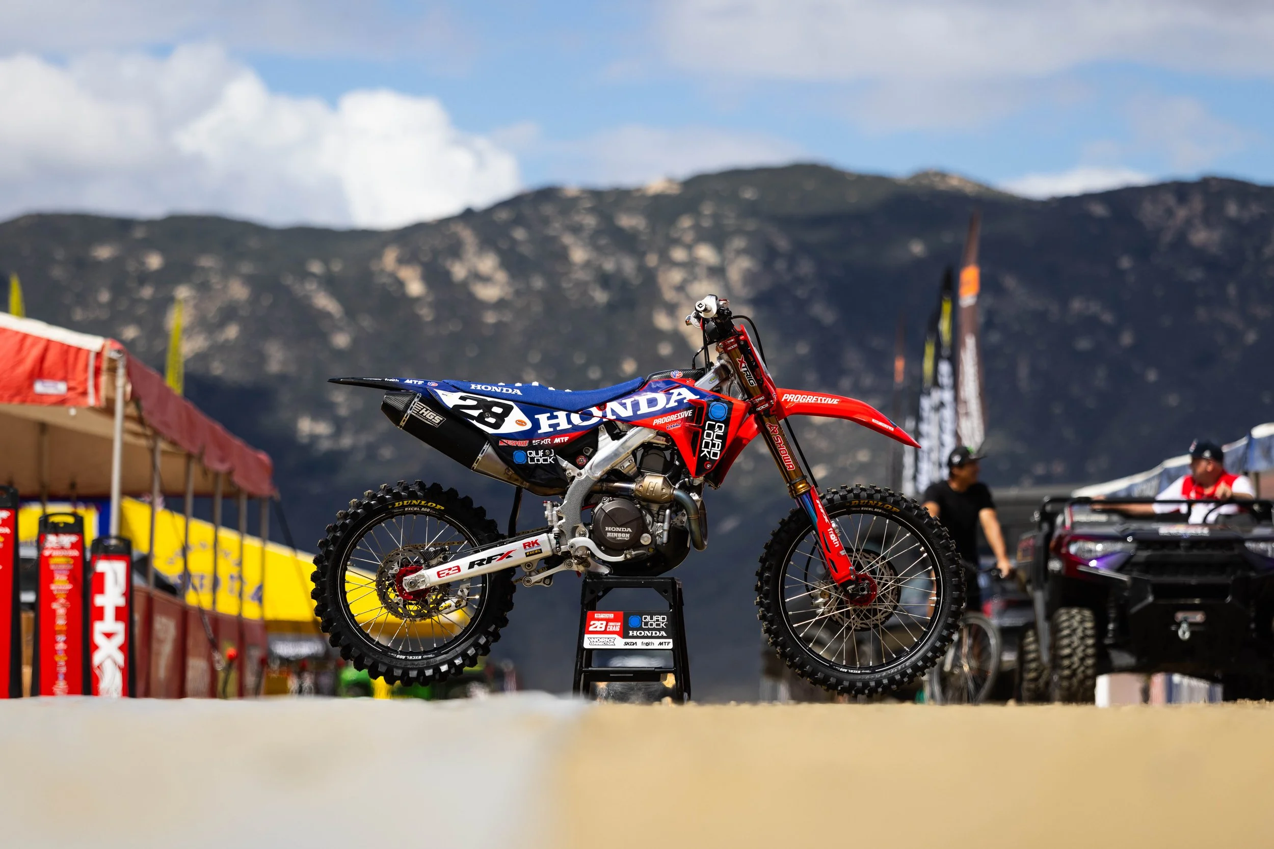 Motocross dirt bike on display at a racing event with mountains in the background, sponsors' banners, and people in the distance.