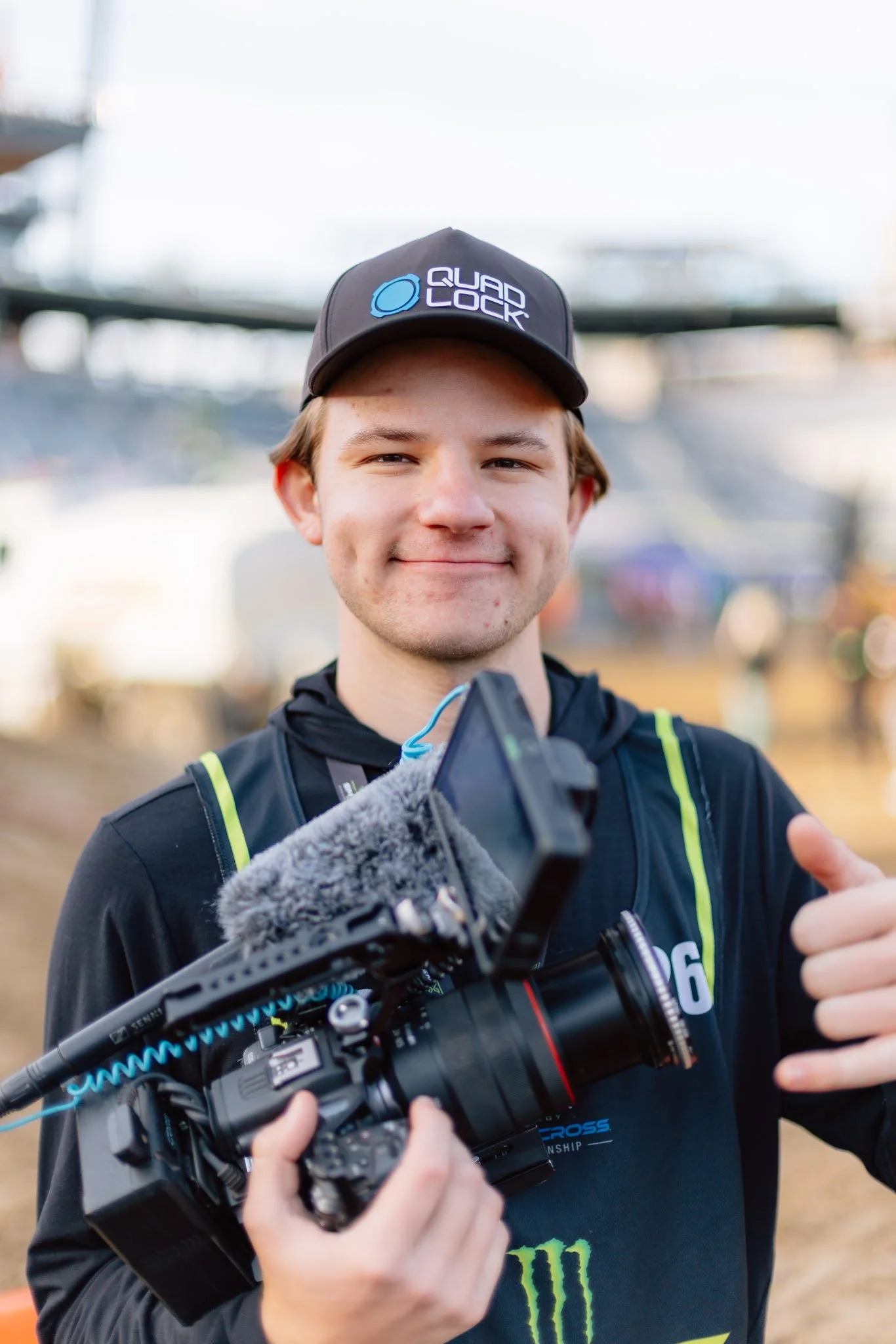 A young man with light brown hair and a slight smile, wearing a cap with a logo that says 'Quad Lock' and a black athletic jacket with green accents, is holding a professional camera with a large lens and microphone attached, while giving a thumbs-up gesture outdoors.